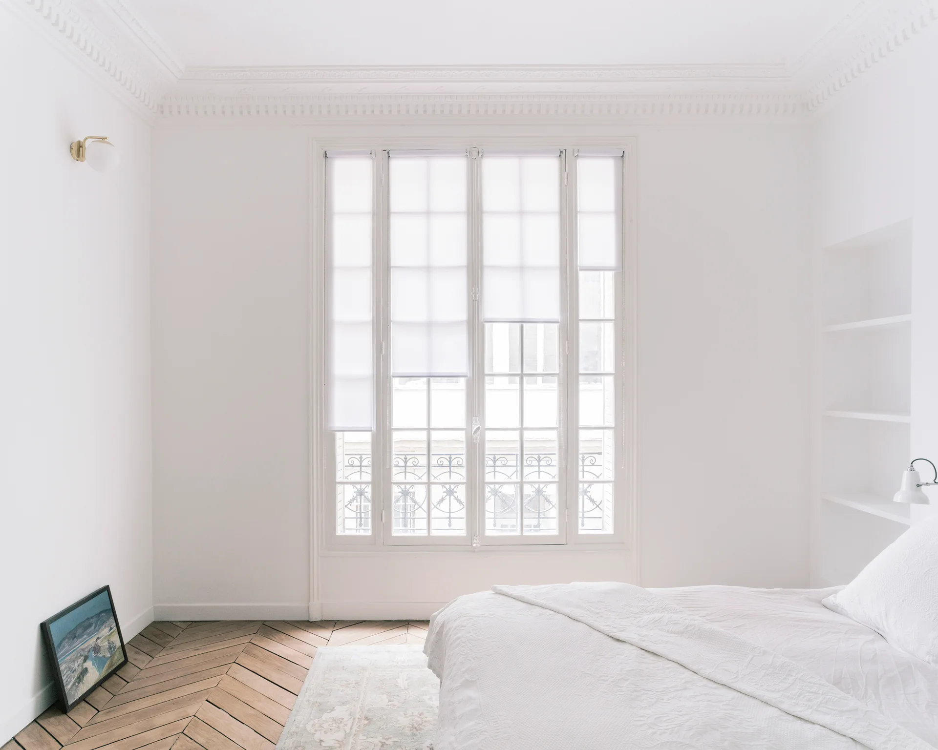 A minimalist white bedroom with a large French window, sheer white roller shades, and light-toned herringbone wood floors.