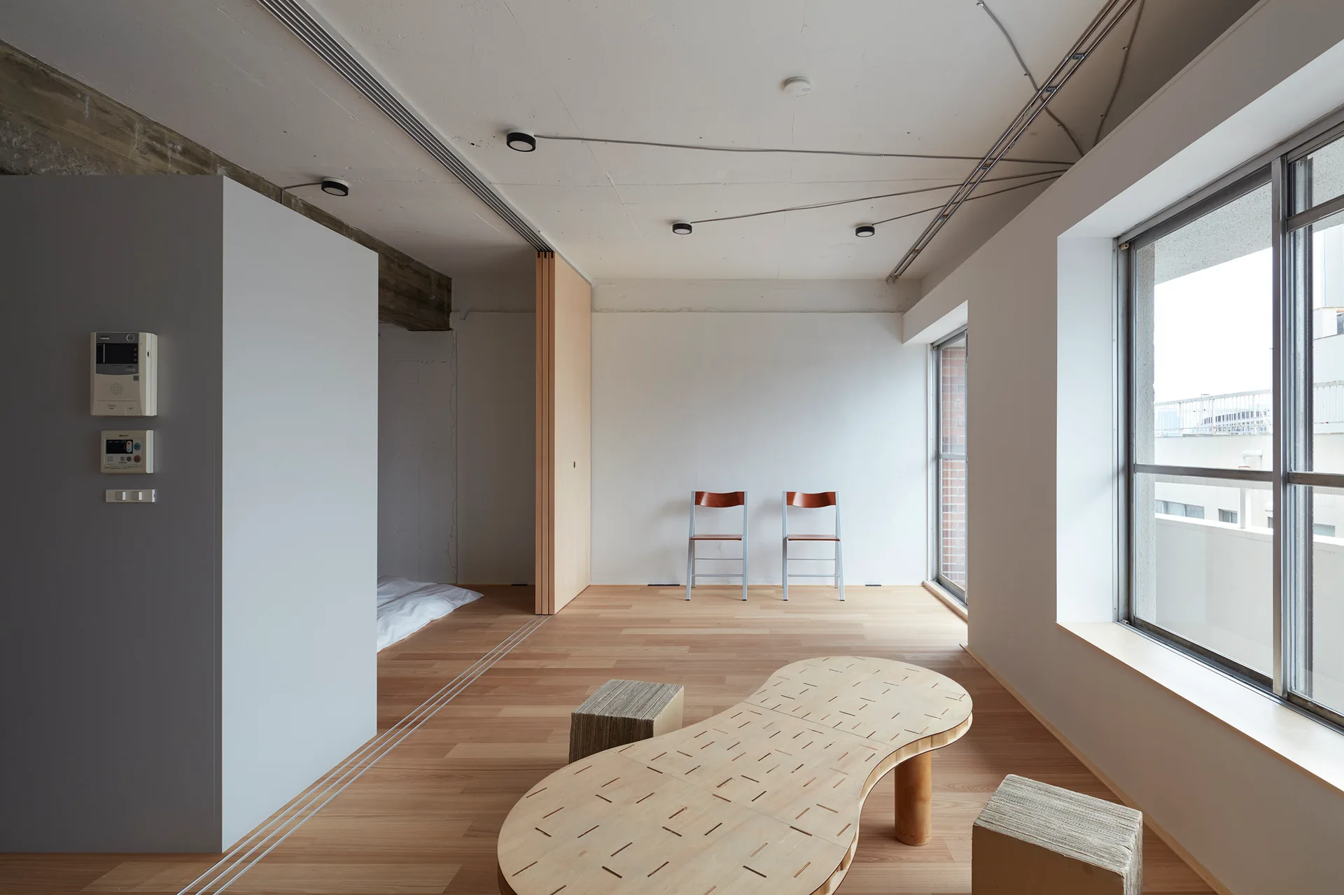 A minimalist living space featuring an organic-shaped wooden coffee table, two simple chairs against a white wall, and a wooden sliding partition.