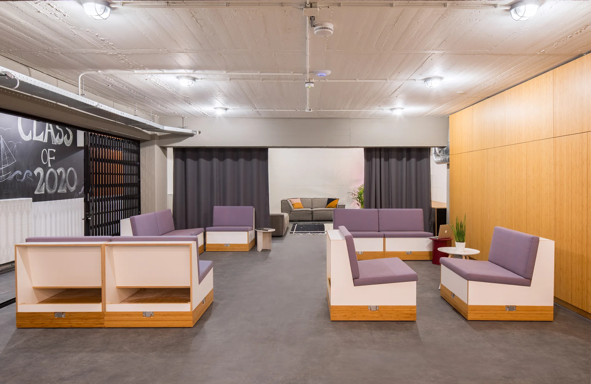 A spacious industrial lounge features purple modular sofas on a grey concrete floor, a light wood-paneled wall, and a textured concrete ceiling.
