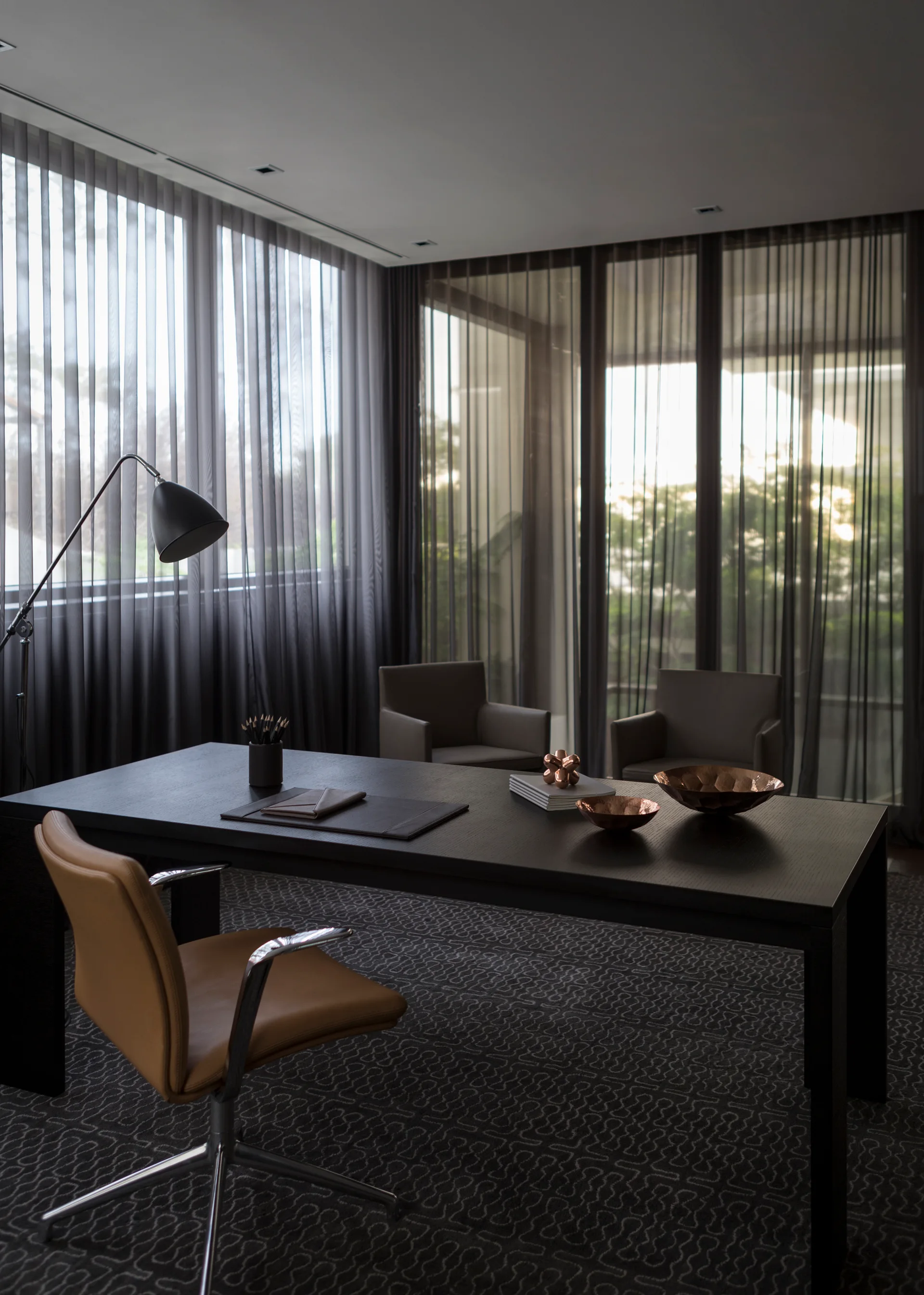 A modern home office features a black wooden desk, a tan leather chair, and floor-to-ceiling windows with dark sheer curtains.
