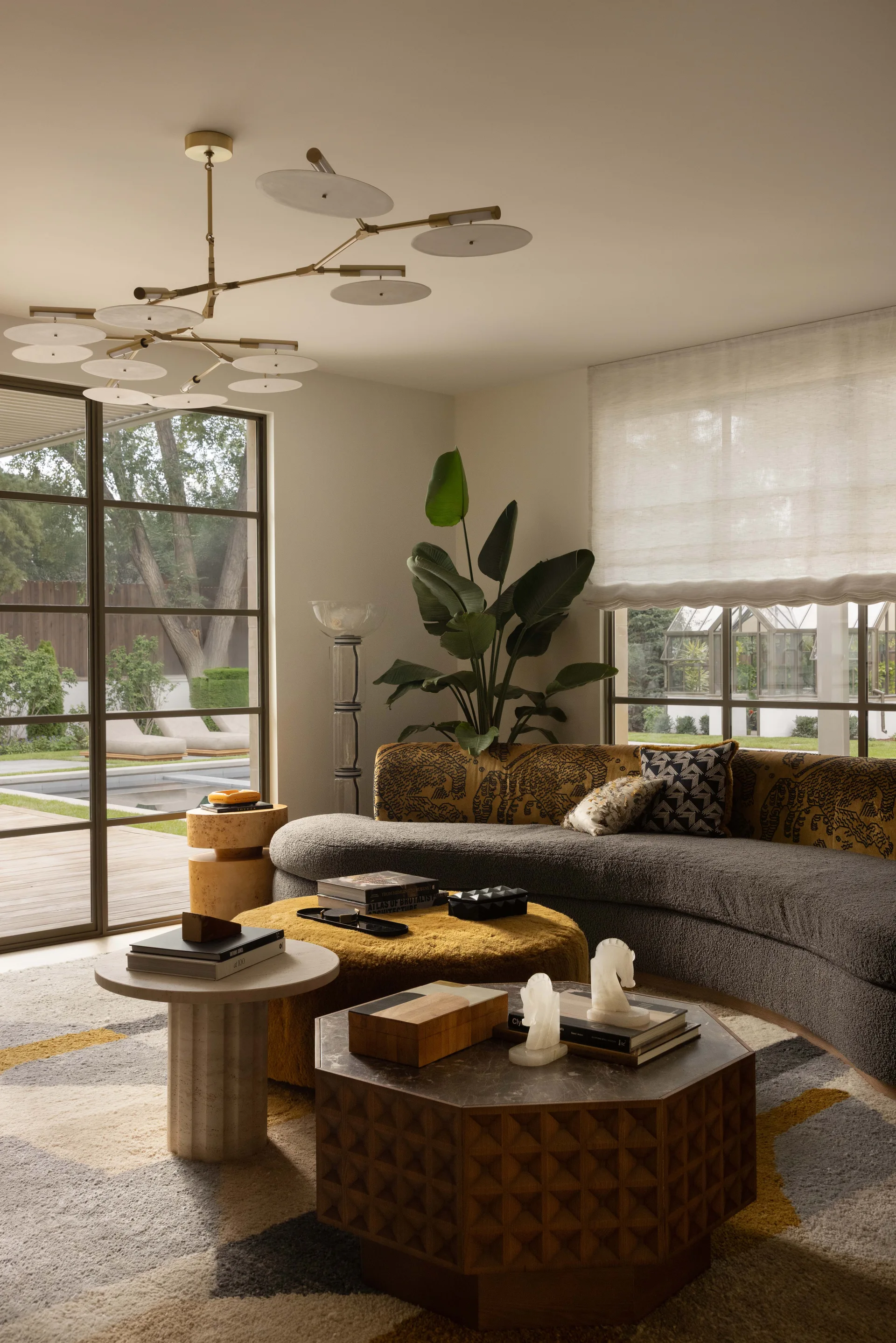 An eclectic living room featuring a curved gray sofa, a mustard yellow ottoman, a hexagonal coffee table, and a large bird of paradise plant.
