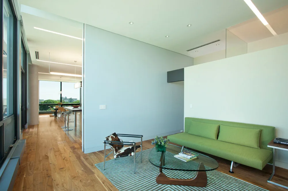 A modern open plan living room features a green sofa, a glass-top coffee table, and a cowhide accent chair on a striped rug.
