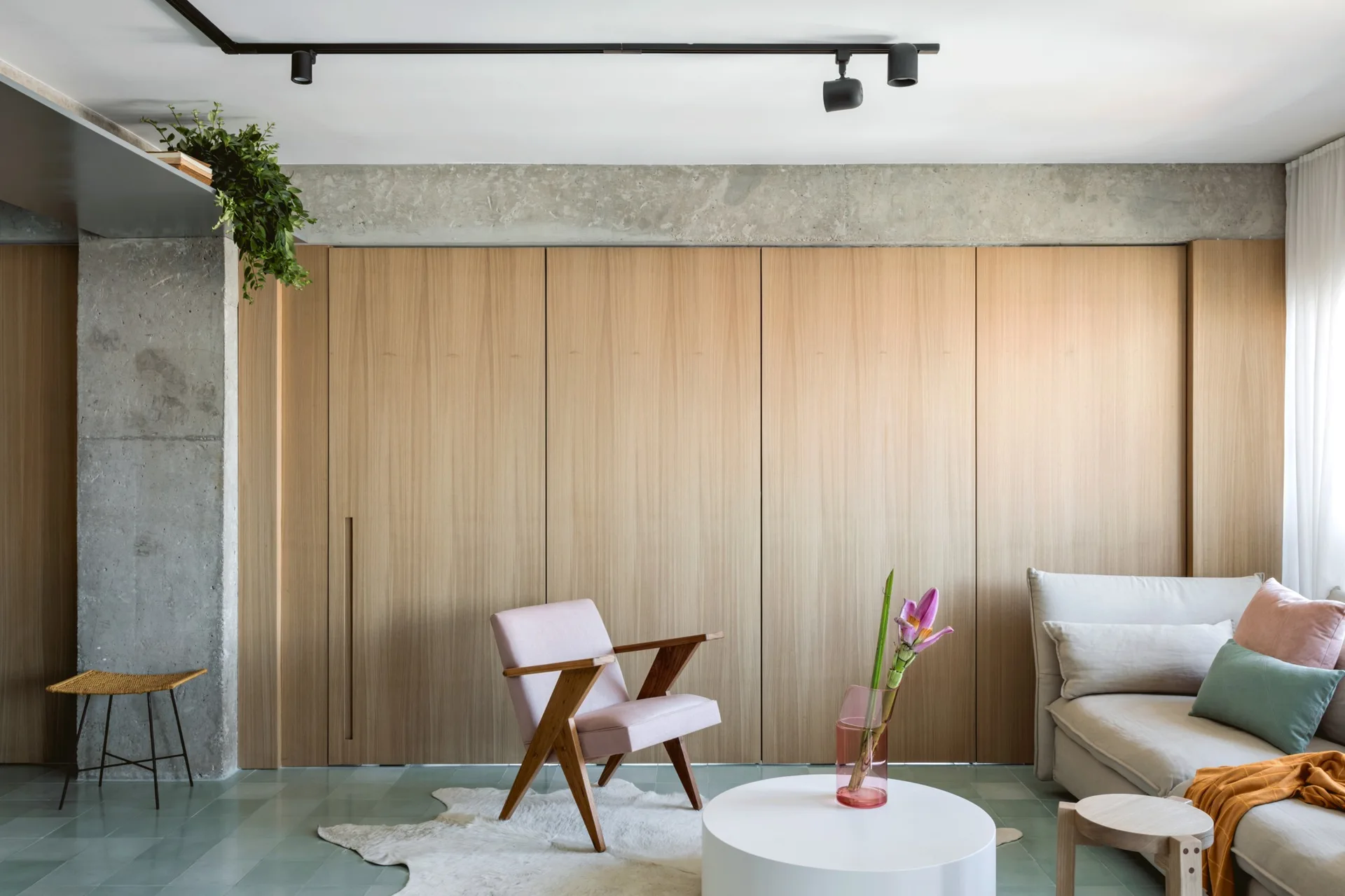 A living room featuring a light wood paneled wall, a raw concrete column, a pink armchair, and a cream sofa on a green tiled floor.