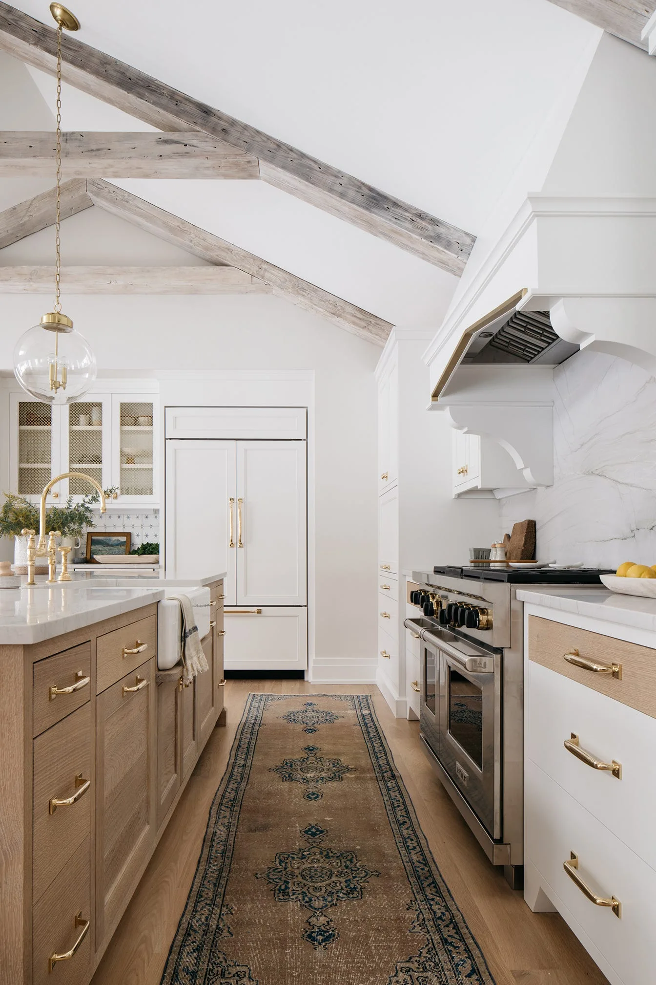 A bright kitchen features white custom cabinetry, a wood-toned island with gold hardware, and rustic exposed ceiling beams.