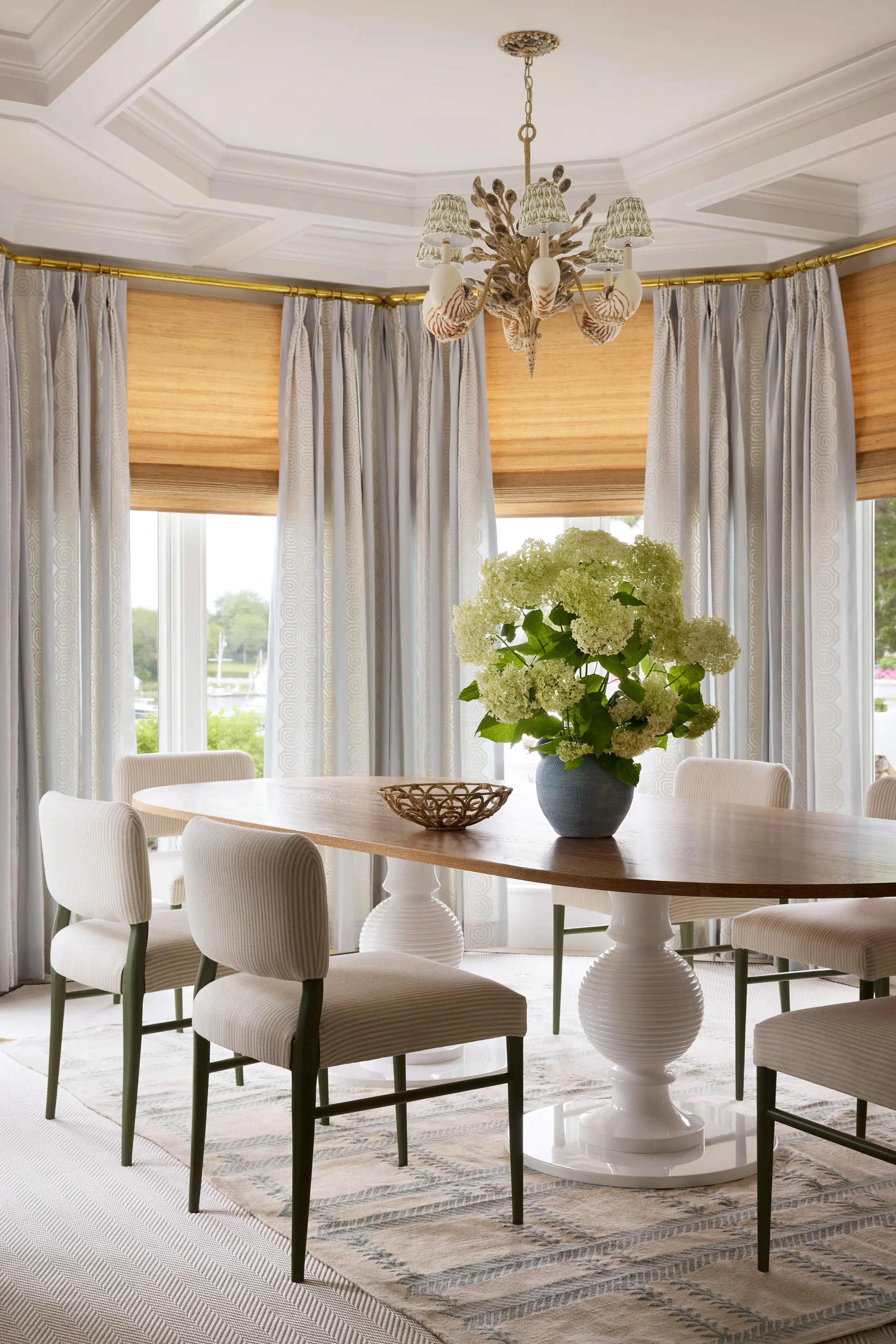 A coastal dining room features an oval wood table with white ribbed pedestal bases, striped dining chairs, and patterned blue drapes.