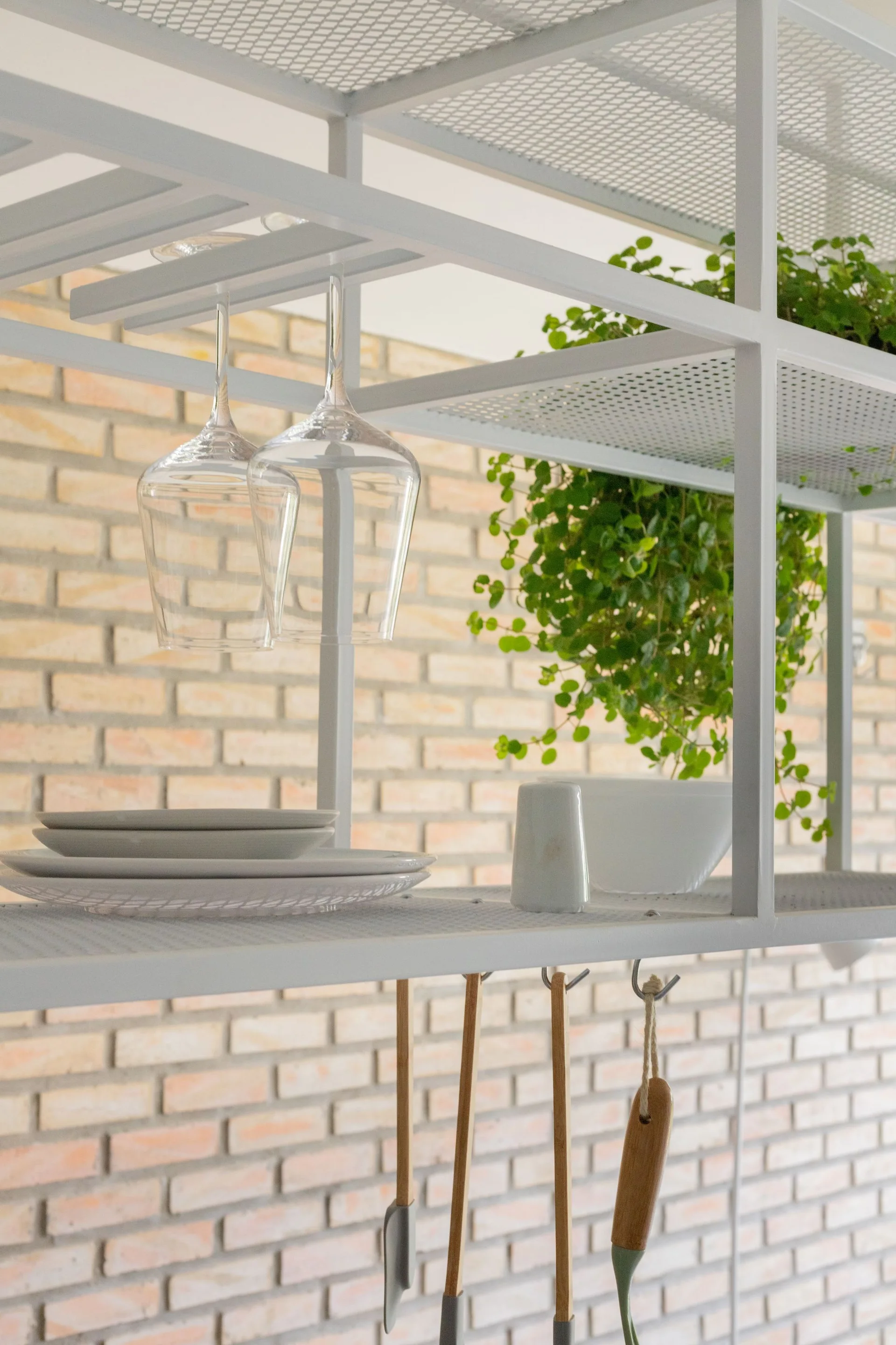 A white metal open shelving unit holds ceramic plates, upside-down wine glasses, and a potted plant against a light tan brick wall.