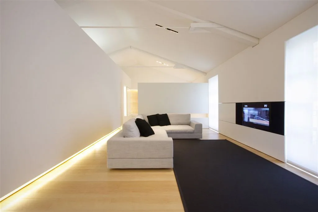 A minimalist white living room featuring a light grey sectional sofa on a black rug, with warm cove lighting along the baseboard.