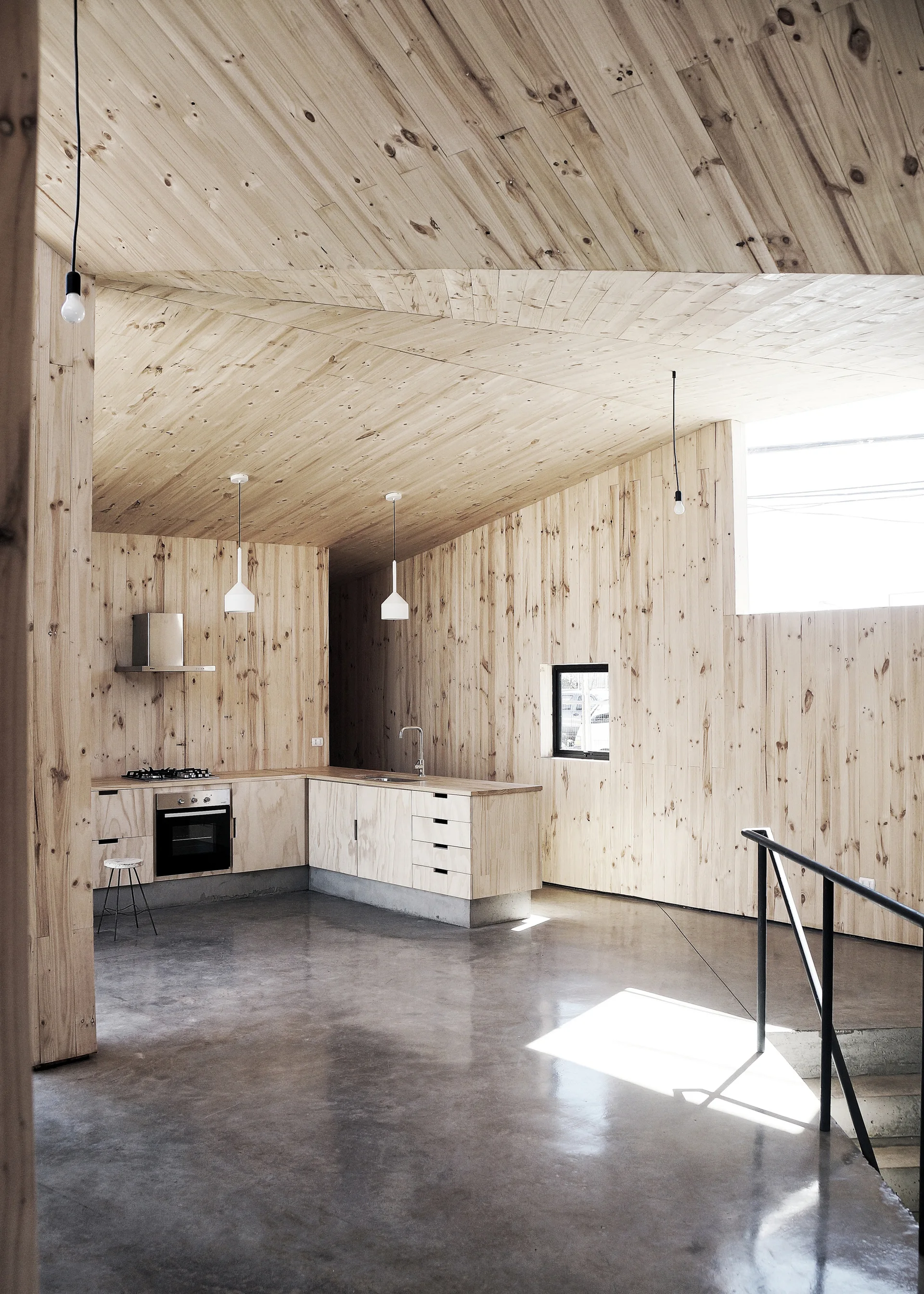 A minimalist kitchen features light wood paneled walls and ceiling, matching cabinetry, a polished concrete floor, and white pendant lights.
