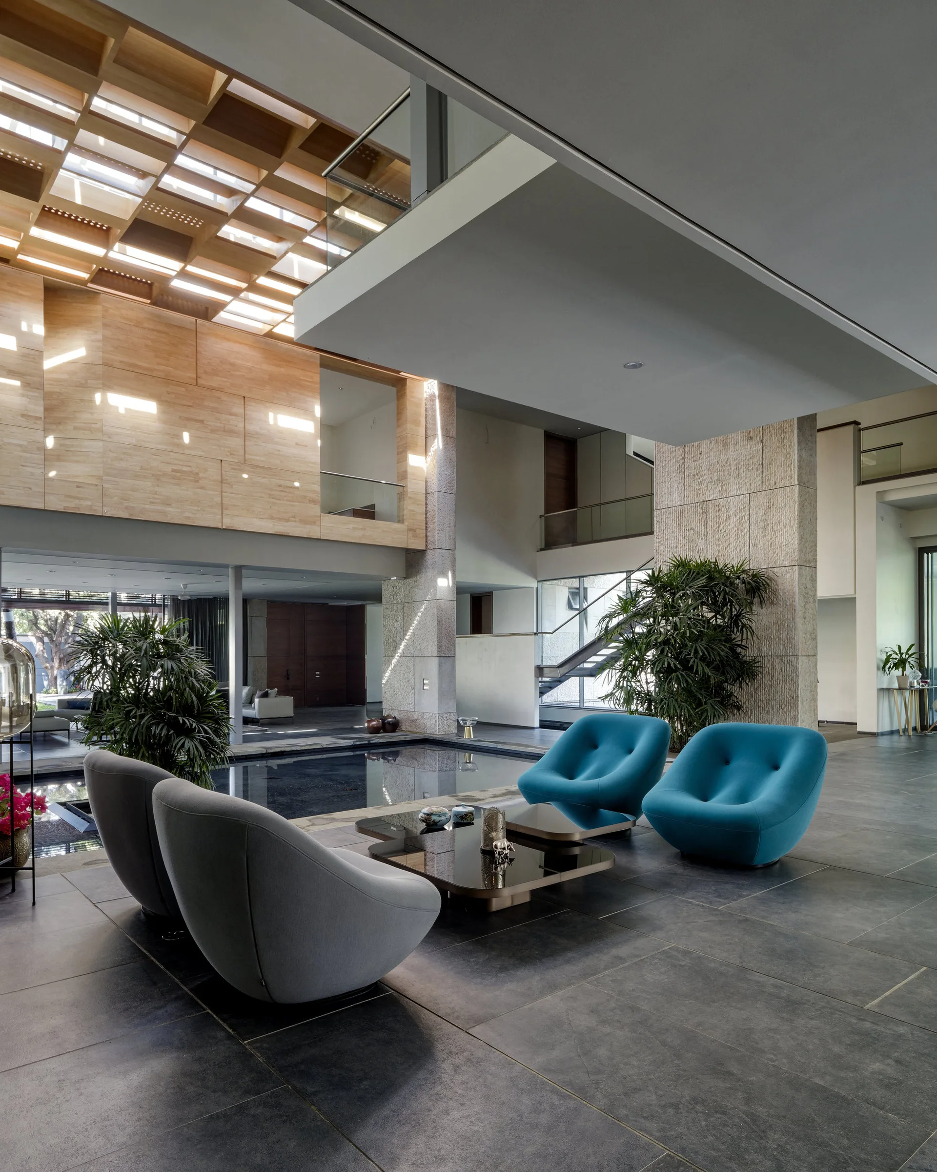 An open-plan living room featuring teal and grey armchairs around a low coffee table next to a sunken indoor pool beneath a wooden grid skylight.