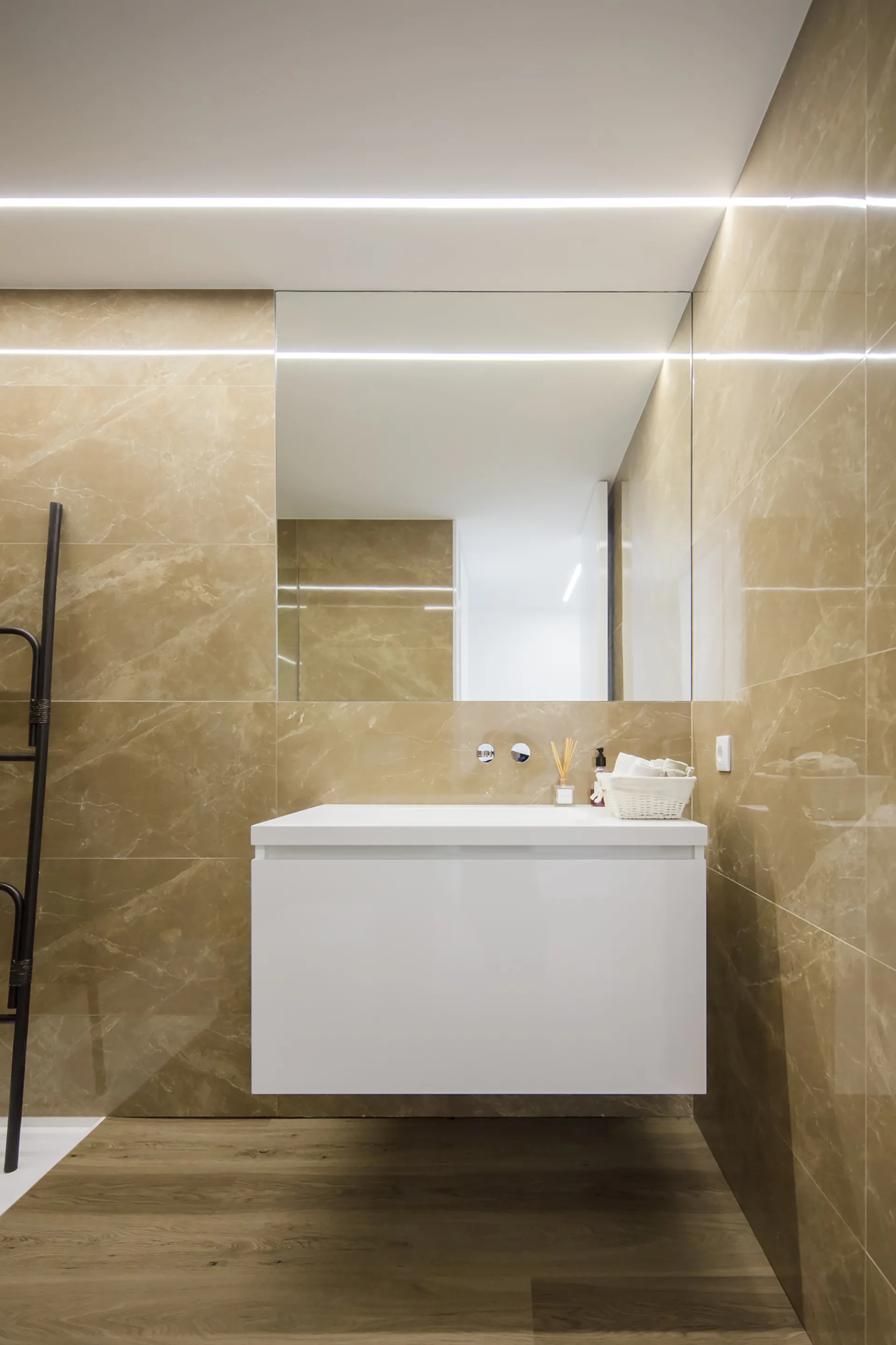 A modern bathroom features a white floating vanity under a large mirror, surrounded by brown marble-patterned wall tiles and wood-look flooring.