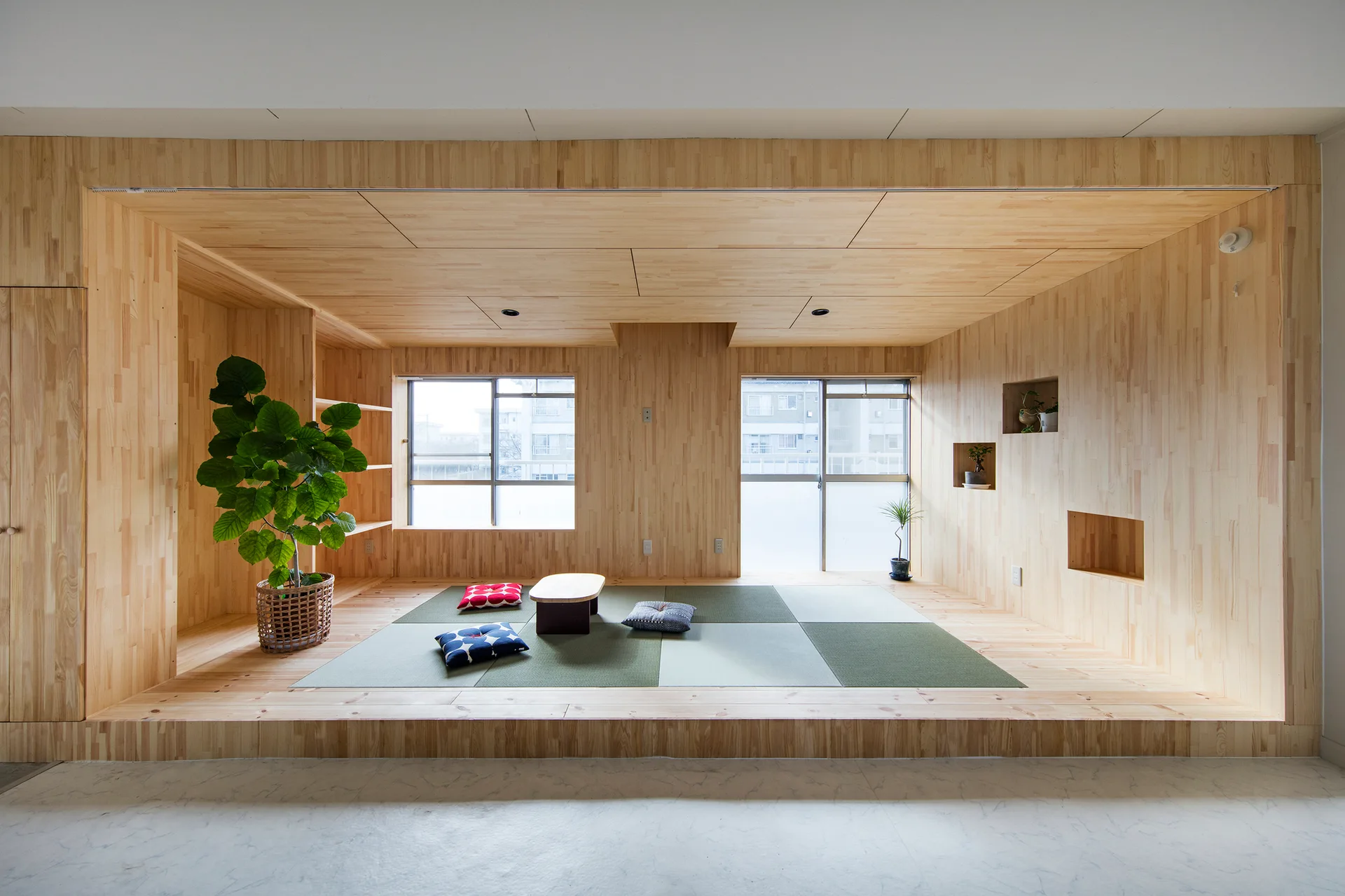 A raised light wood platform with tatami mats, a low table, cushions, and recessed wall niches is enclosed by matching wooden walls and ceiling.