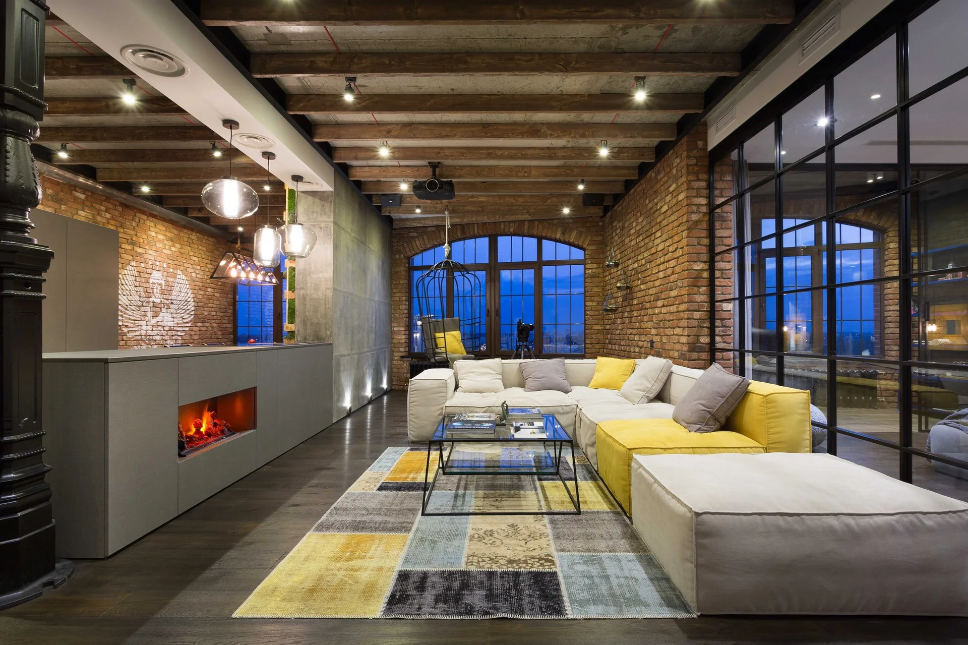 An industrial loft living room with a grey and yellow modular sectional sofa, exposed brick walls, and a wooden beam ceiling.