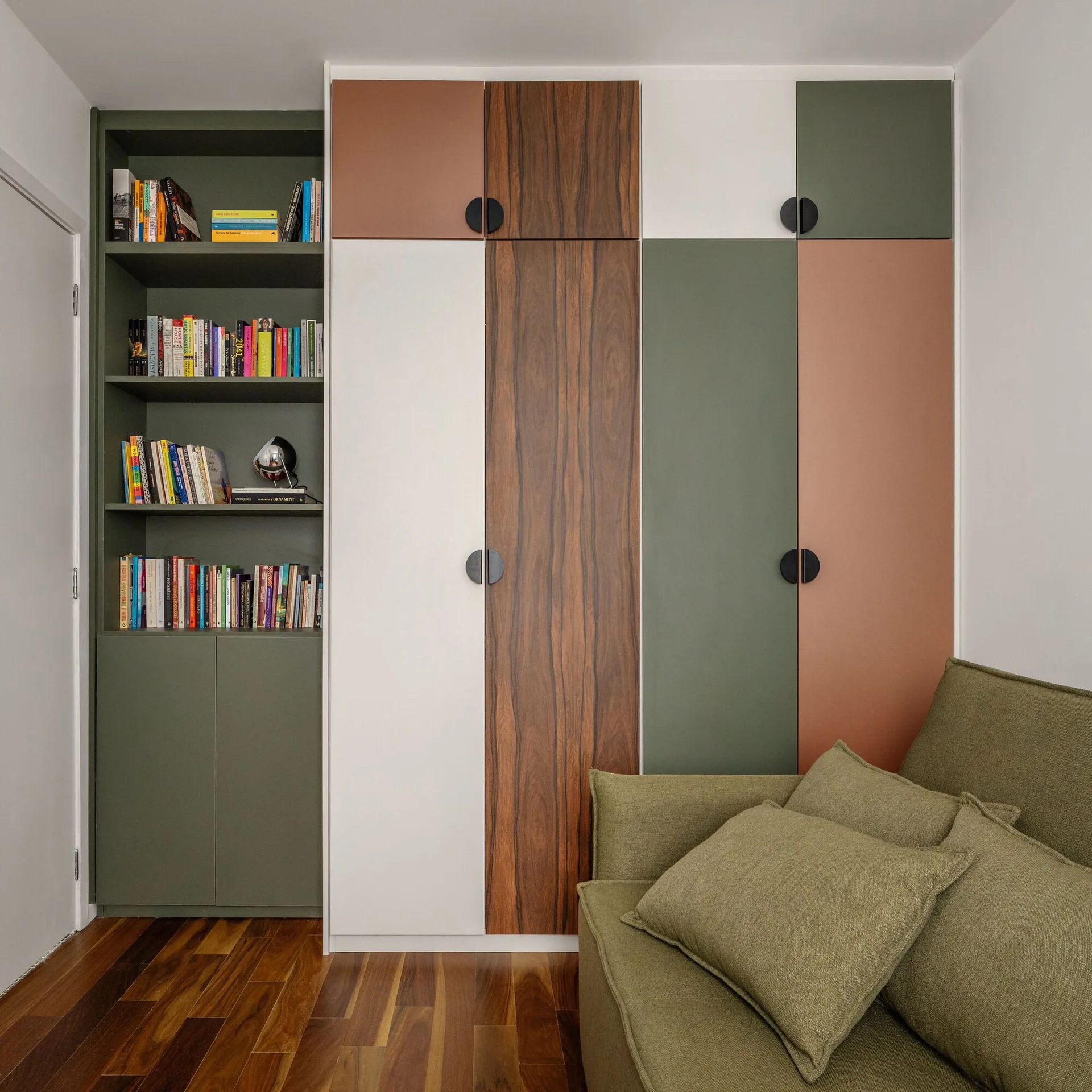 A custom wardrobe featuring terracotta, olive green, and wood panels stands beside an integrated bookshelf and a green fabric sofa on hardwood floors.