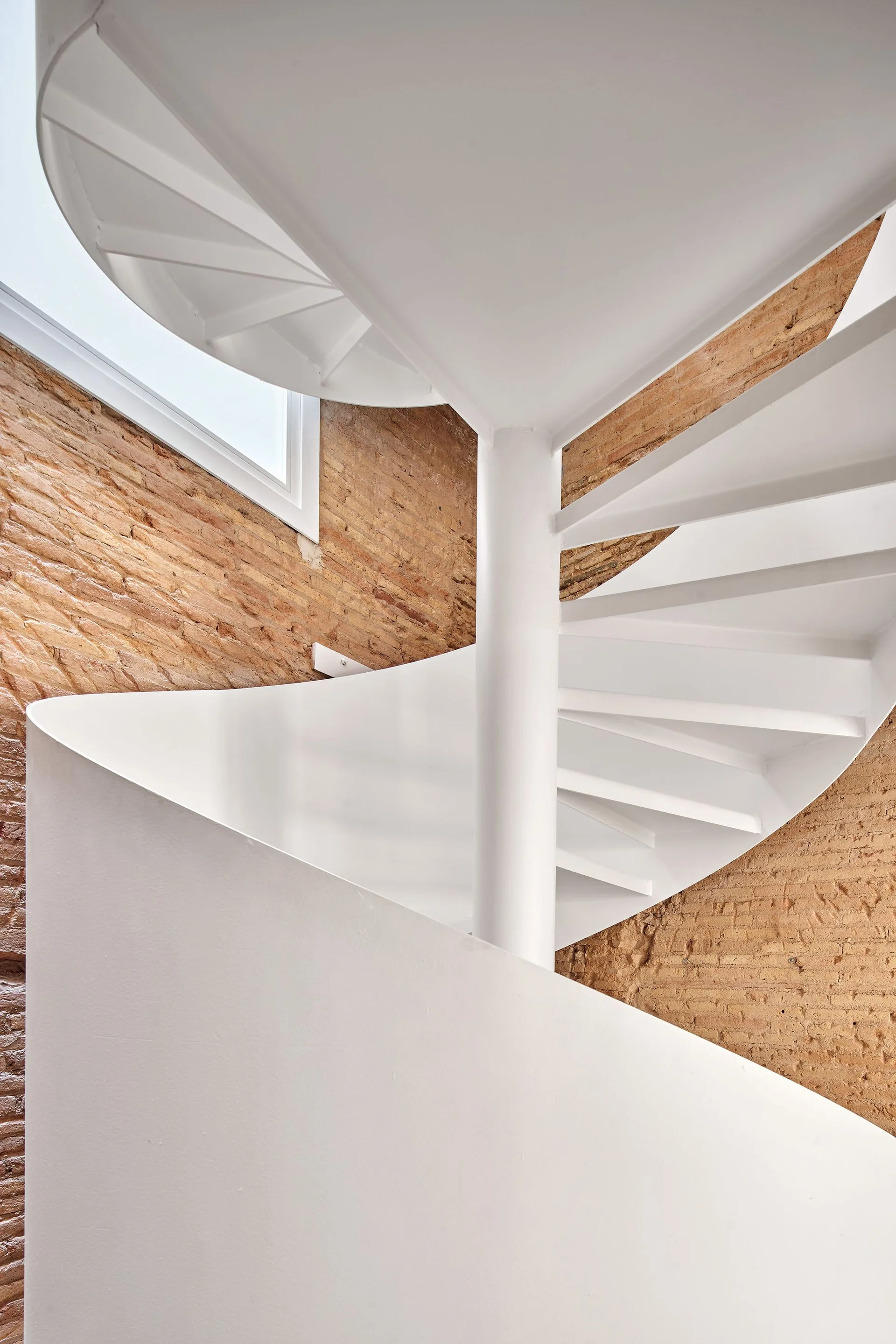 A white metal spiral staircase curves upward in front of a textured red brick wall, with a small window visible near the top.