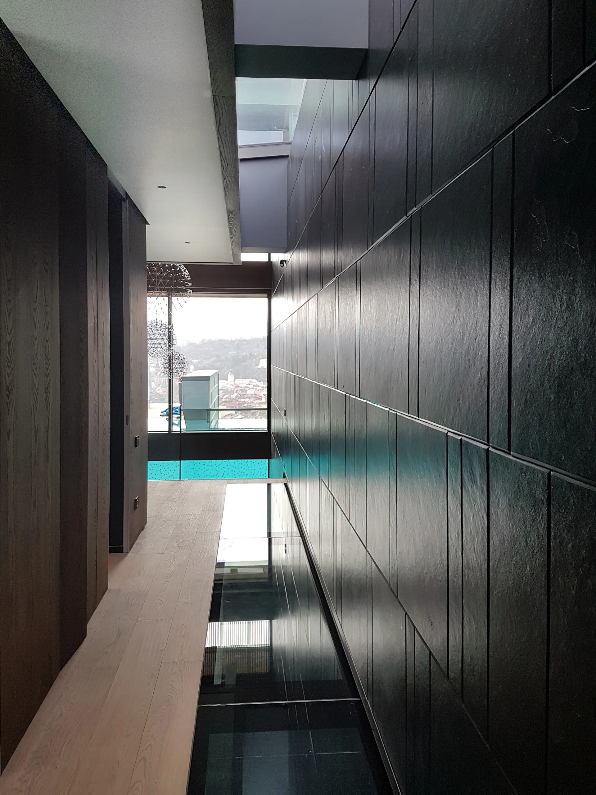 A minimalist modern hallway with dark textured wall panels on one side, light wood flooring, and a long reflective glass floor runway.