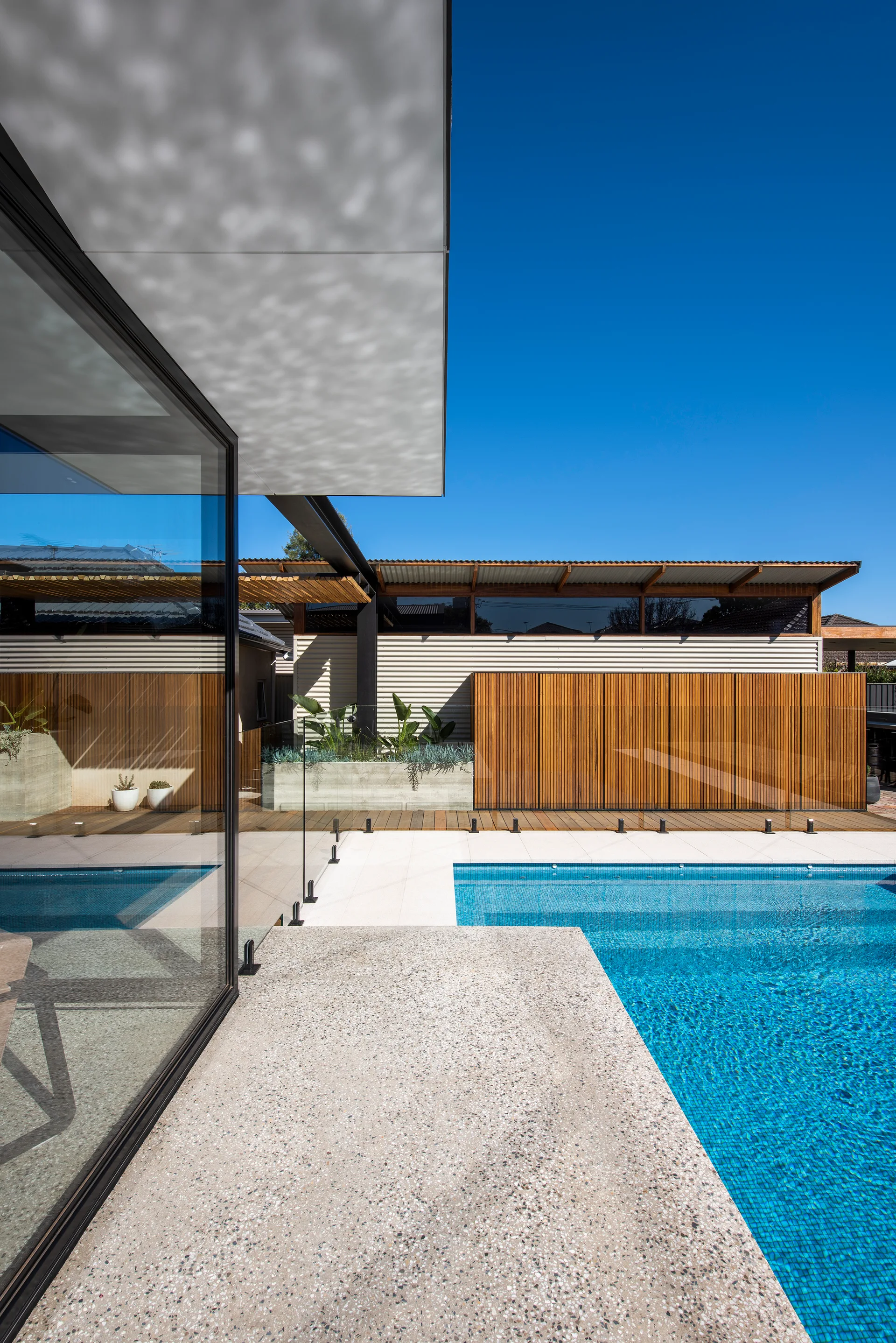 A backyard swimming pool featuring light terrazzo decking, a clear glass safety fence, and a vertical wood slat privacy wall.