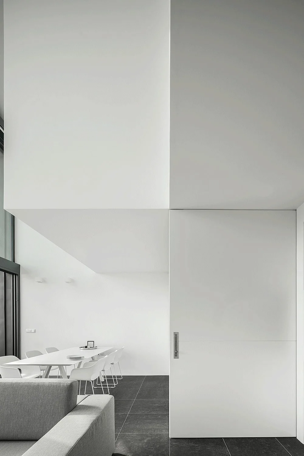 A minimalist living and dining space with a grey sofa, white dining table and chairs, and a large white floor-to-ceiling sliding door on dark tile.
