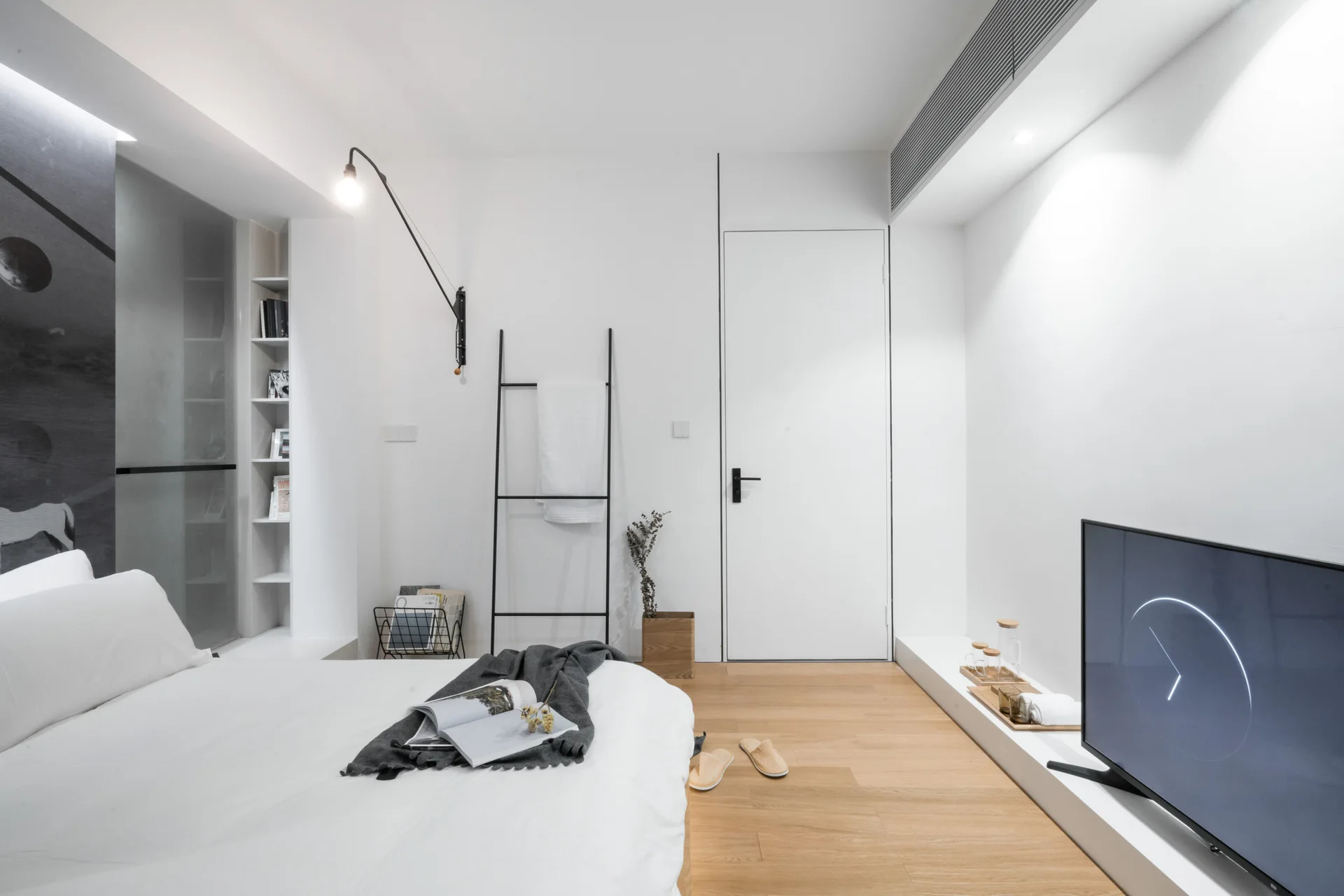 A minimalist white bedroom with light wood flooring, a low platform bed, a black leaning ladder, and a television on a low white plinth.