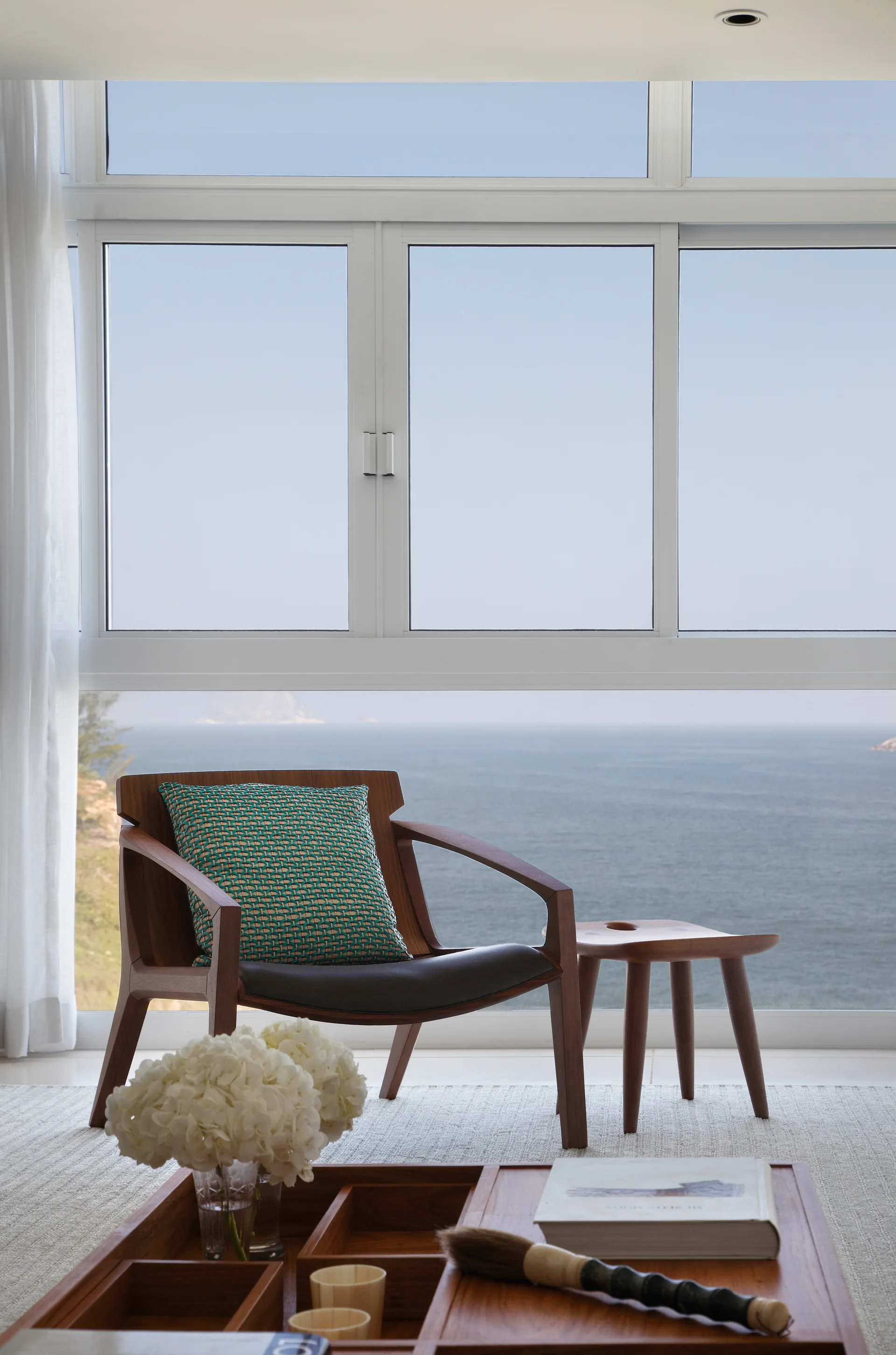 A wooden armchair with a teal patterned pillow and a matching stool sit on a textured rug in front of a large window overlooking the ocean.