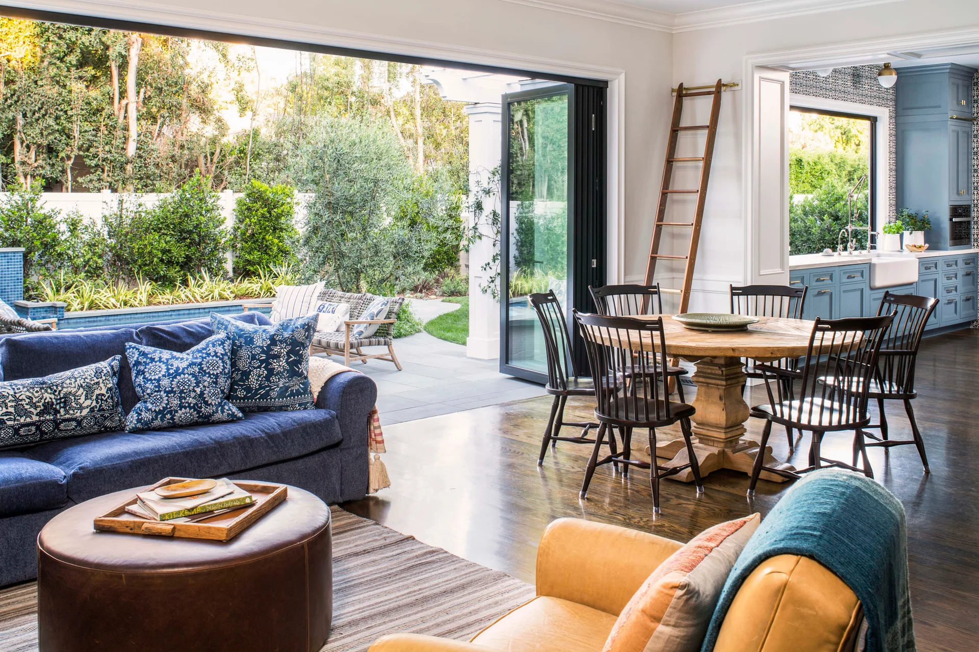 An open-plan living room features a blue sofa, a round wooden dining table with spindle chairs, and large folding glass doors that open to a patio.