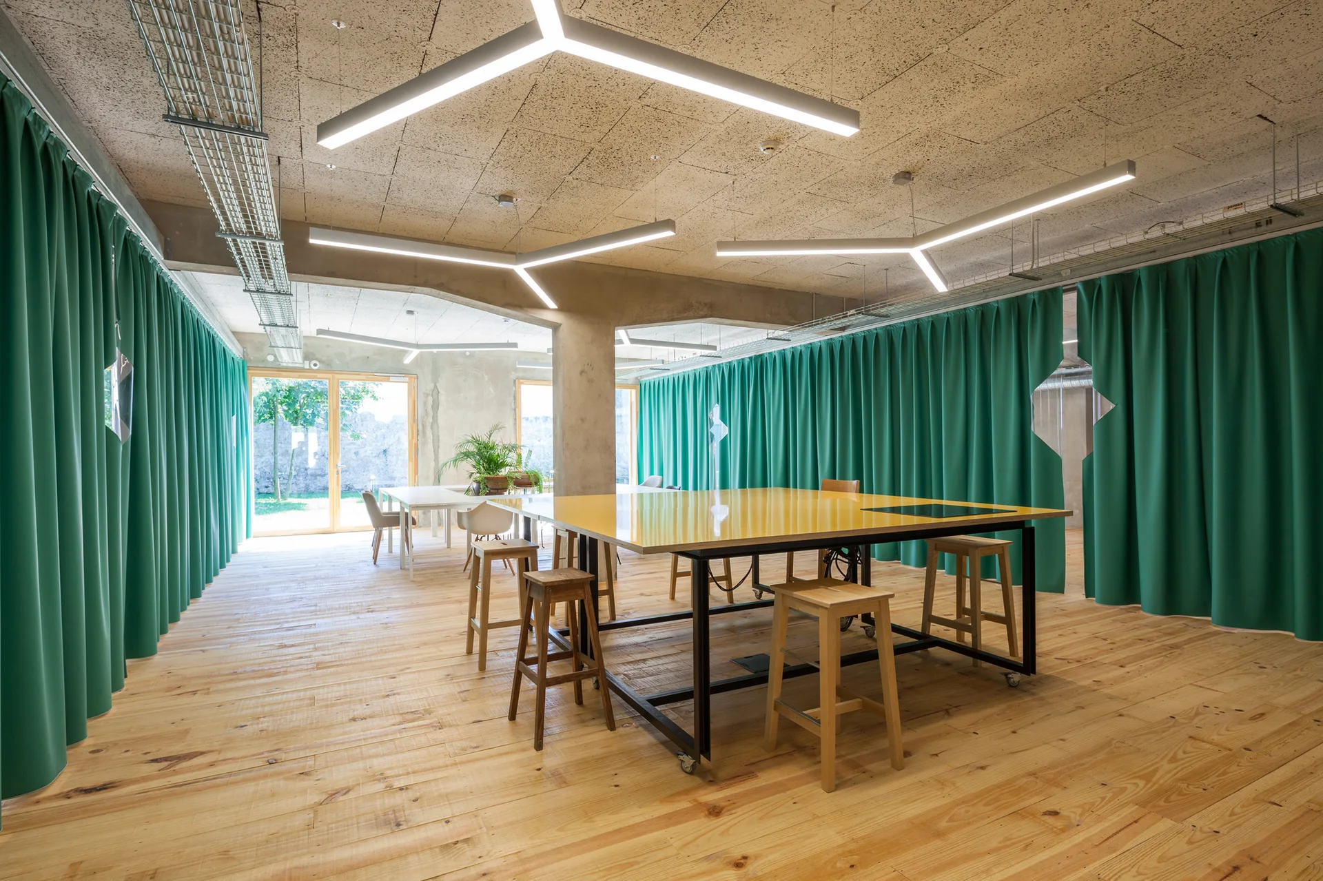 A spacious creative studio features a large yellow work table surrounded by wooden stools, wood flooring, and green curtains covering the walls.