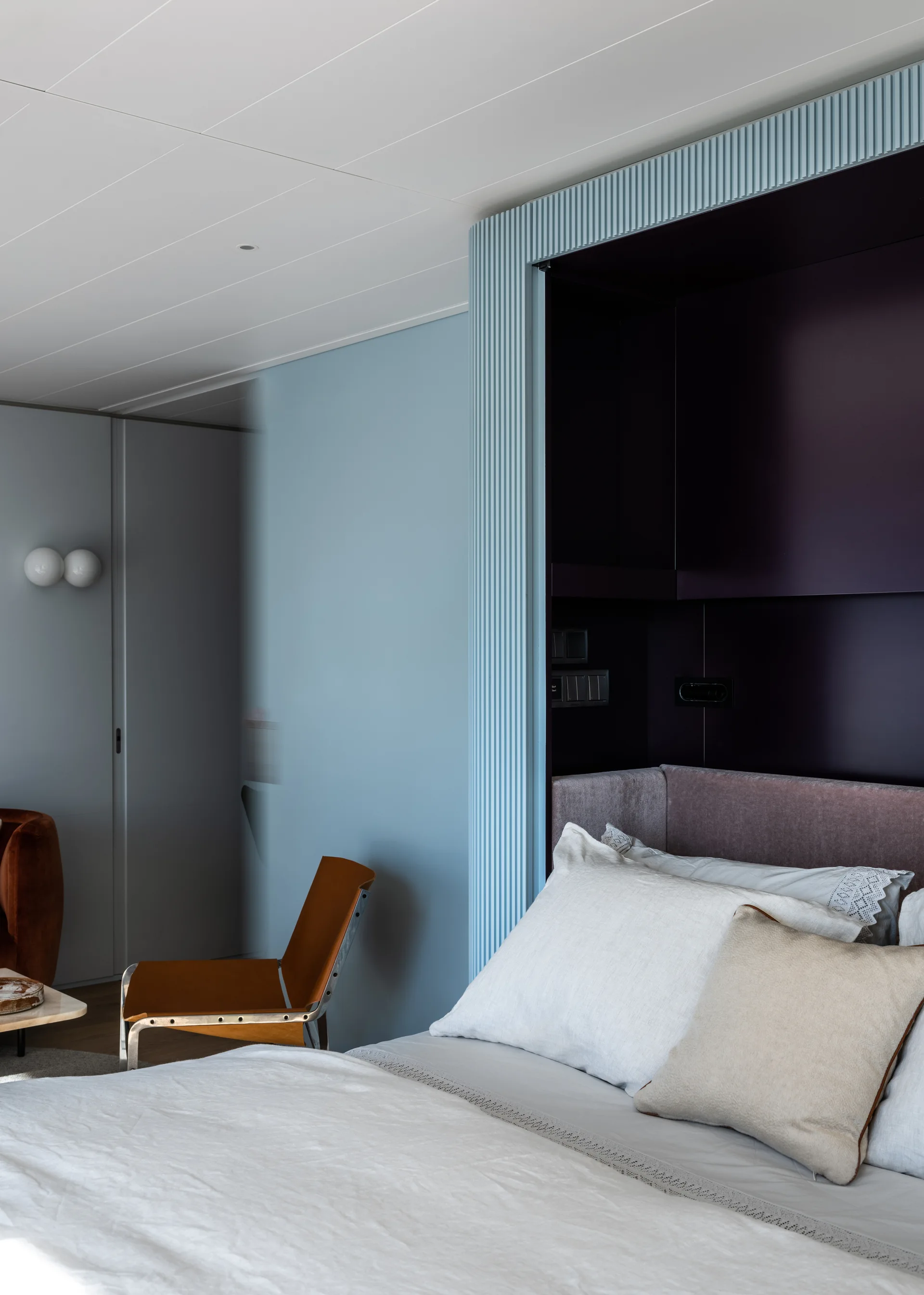 A modern bedroom features a light blue fluted wall framing a deep purple headboard niche next to white bedding and a brown leather chair.