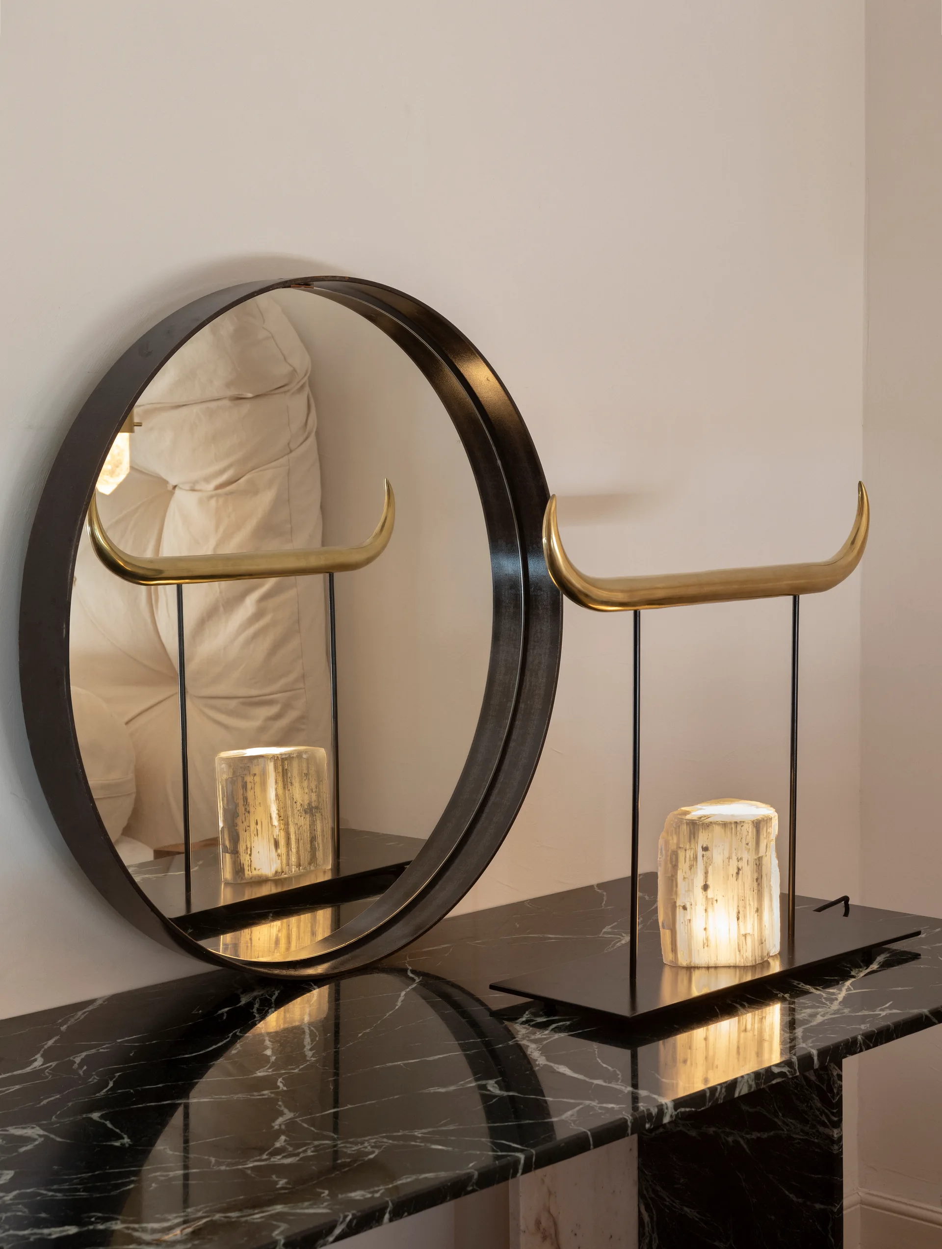 A close-up of a black veined marble console table with a round black mirror and a sculptural brass horn lamp featuring a glowing stone base.