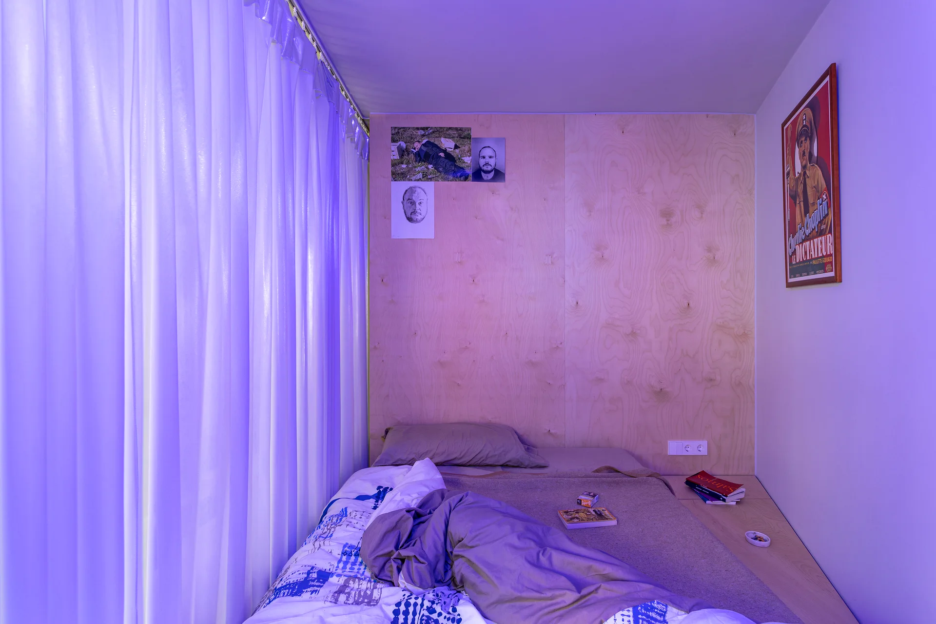 A low platform bed is positioned against a raw plywood wall in a compact bedroom illuminated by purple ambient light.