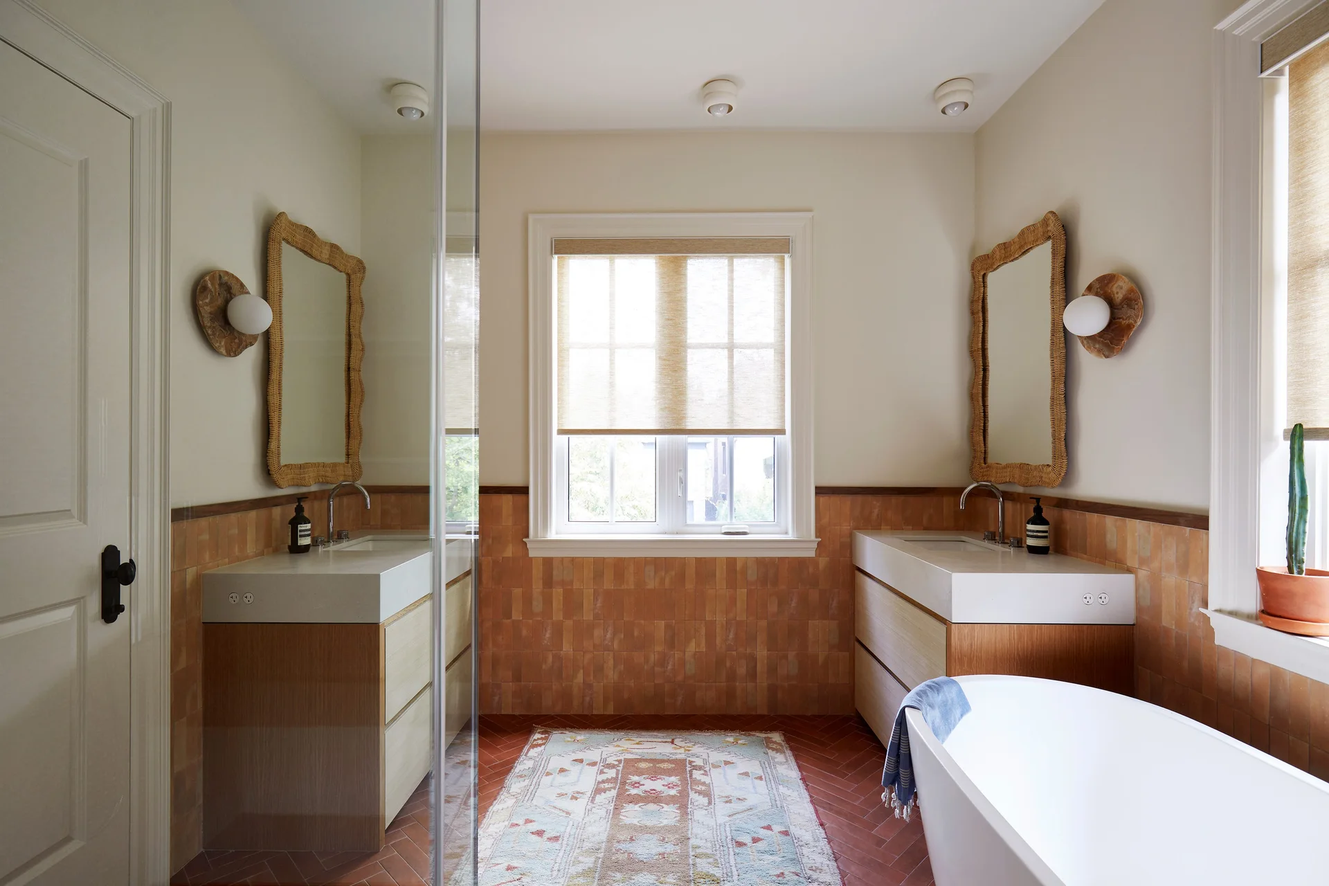 A symmetrical bathroom featuring dual wood vanities, matching woven mirrors, terracotta wall tiles, herringbone floors, and a freestanding white bathtub.
