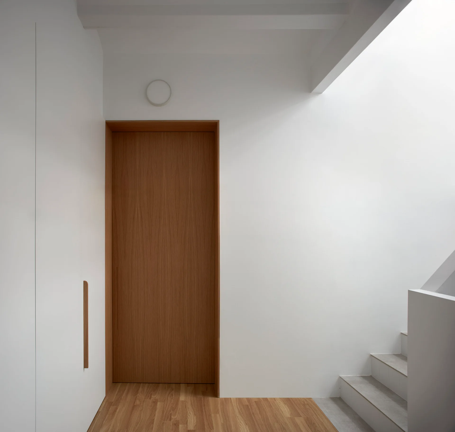 A minimalist white hallway features a central natural wood door, seamless white built-in closets with a wooden handle, and a staircase.