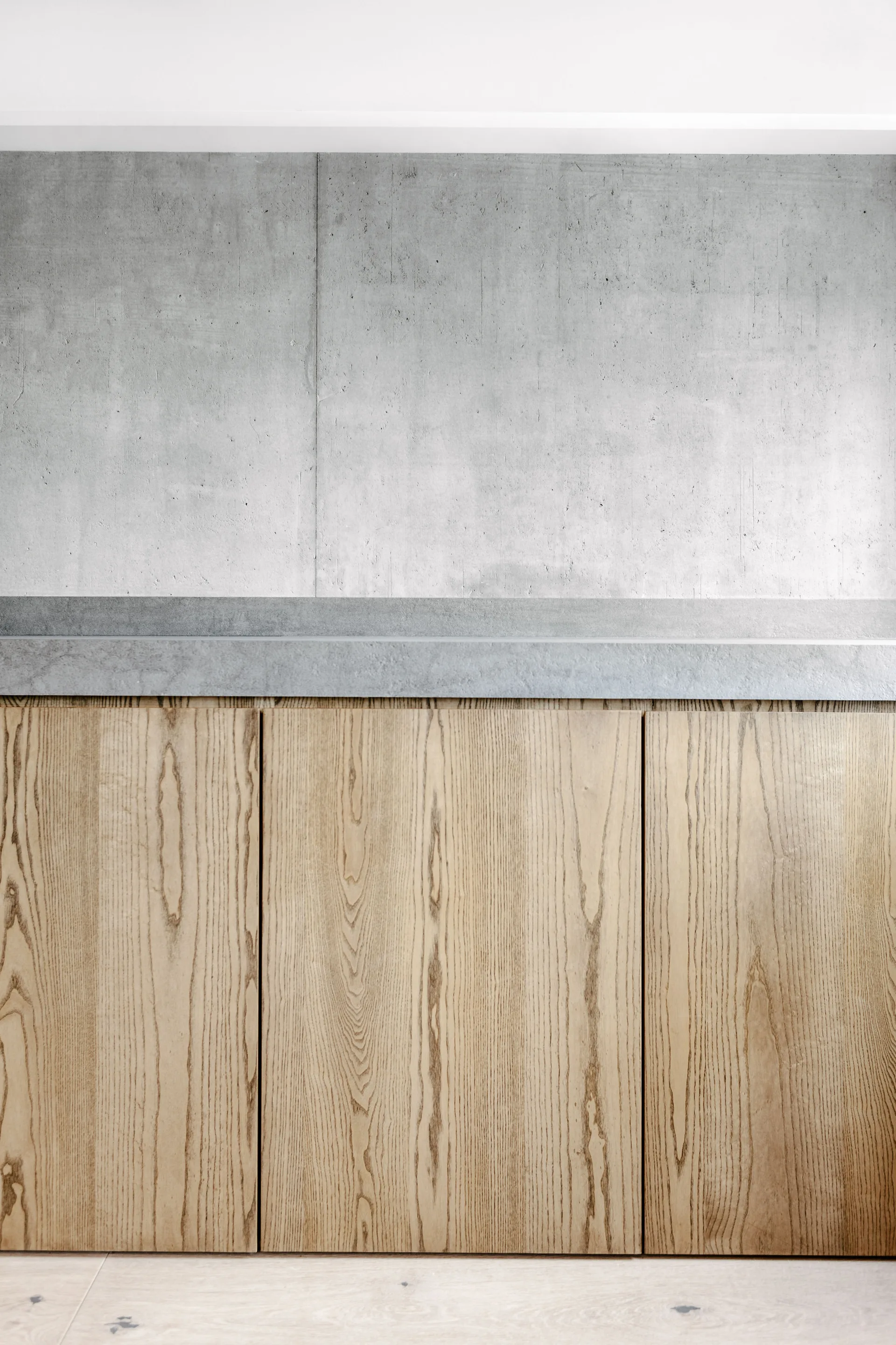 A close-up view of minimalist wood cabinets with visible grain below a slim grey stone countertop and a concrete-textured wall.