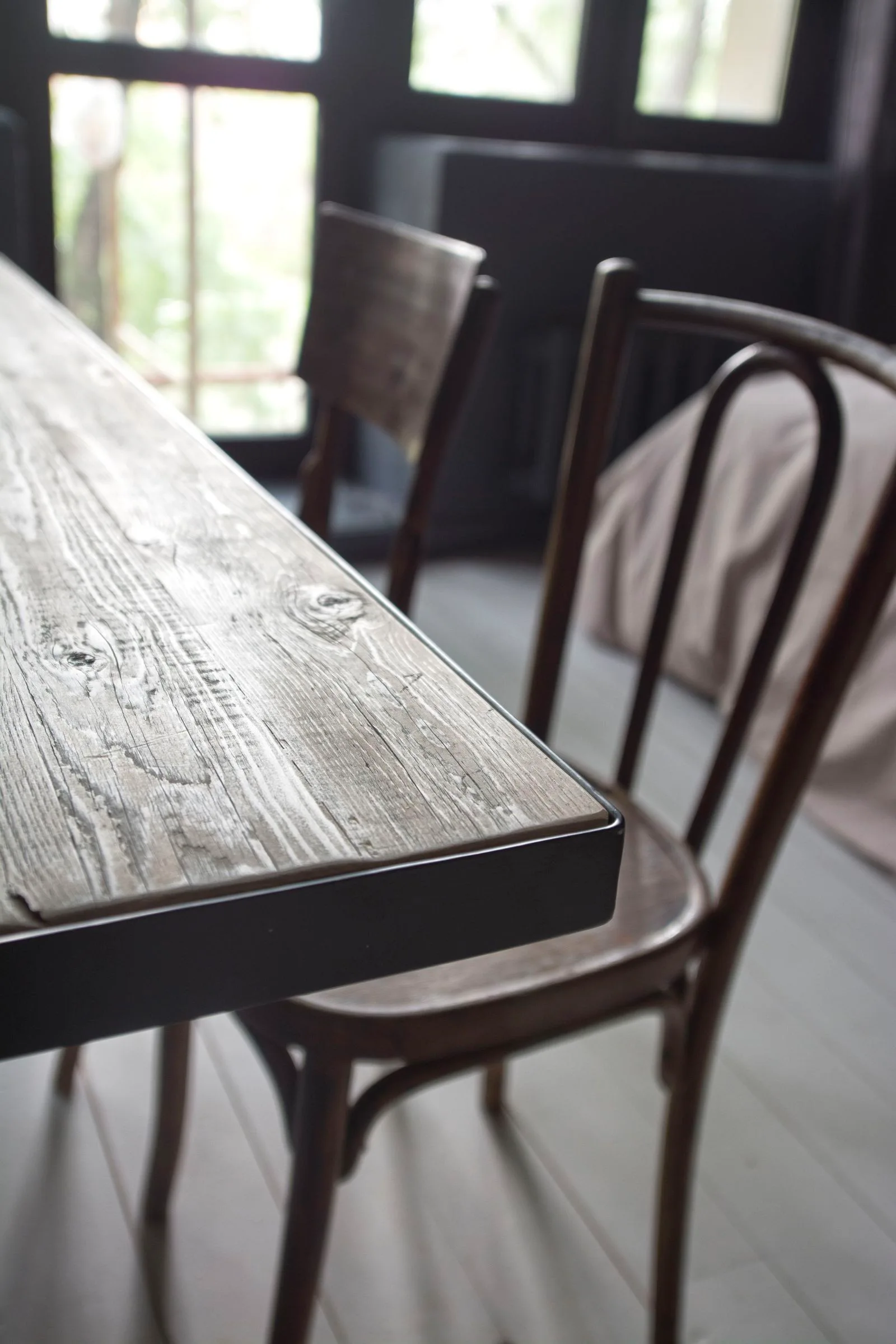 A close-up of a rustic wooden dining table with a dark metal edge, with dark bentwood chairs in a dimly lit room.