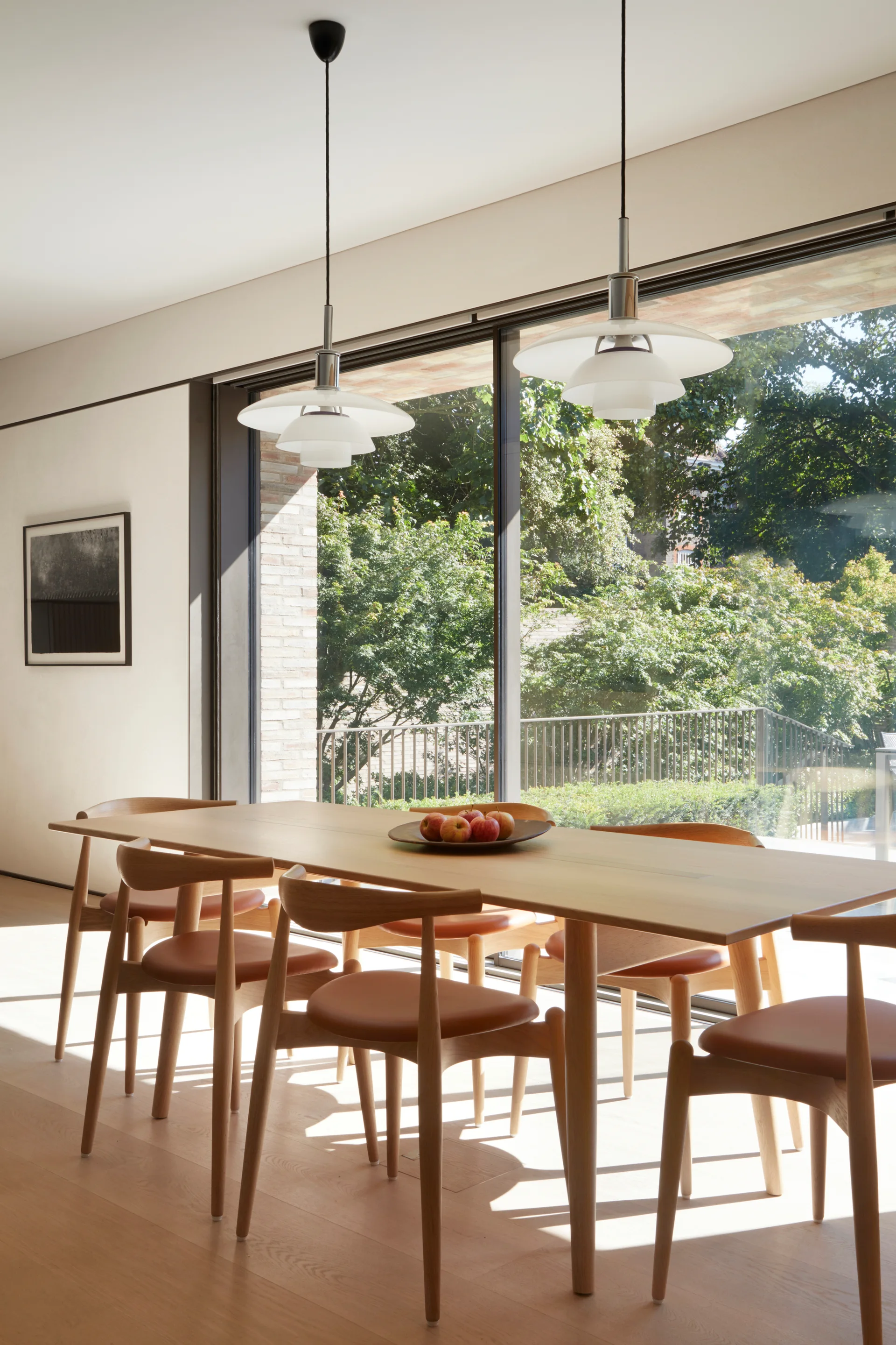 A light wood dining table and matching chairs sit on a hardwood floor beneath two white pendant lights in a sunlit dining room next to a large window.