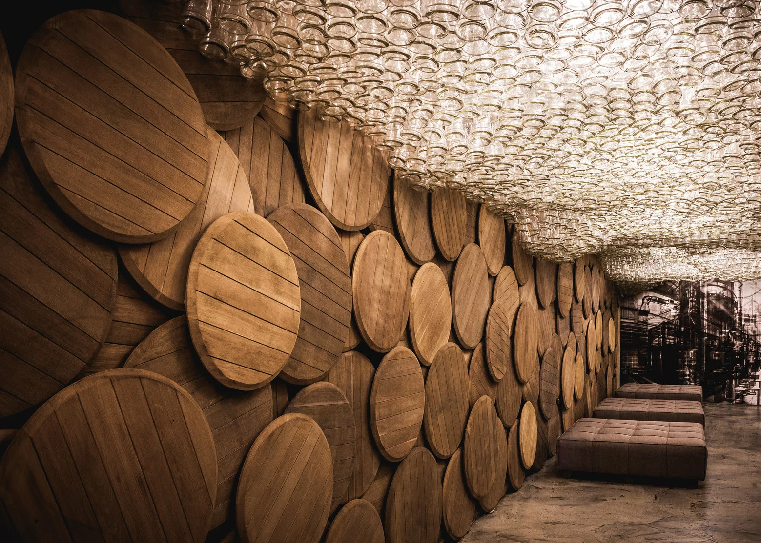 Overlapping circular wooden barrel lids line the wall of a lounge beneath a ceiling canopy made of clear glass bottle bottoms.