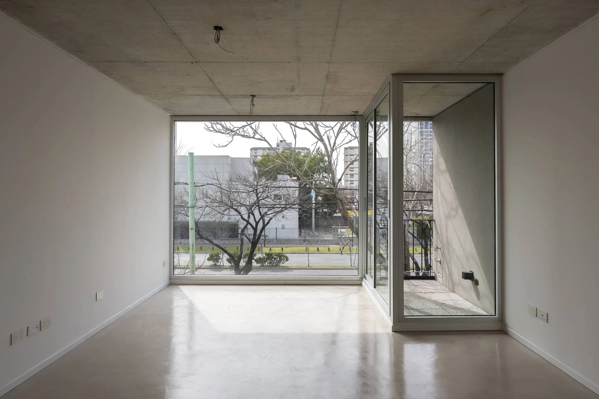 An empty minimalist room with white walls, a smooth light grey floor, a raw concrete ceiling, and glass windows leading to a balcony.