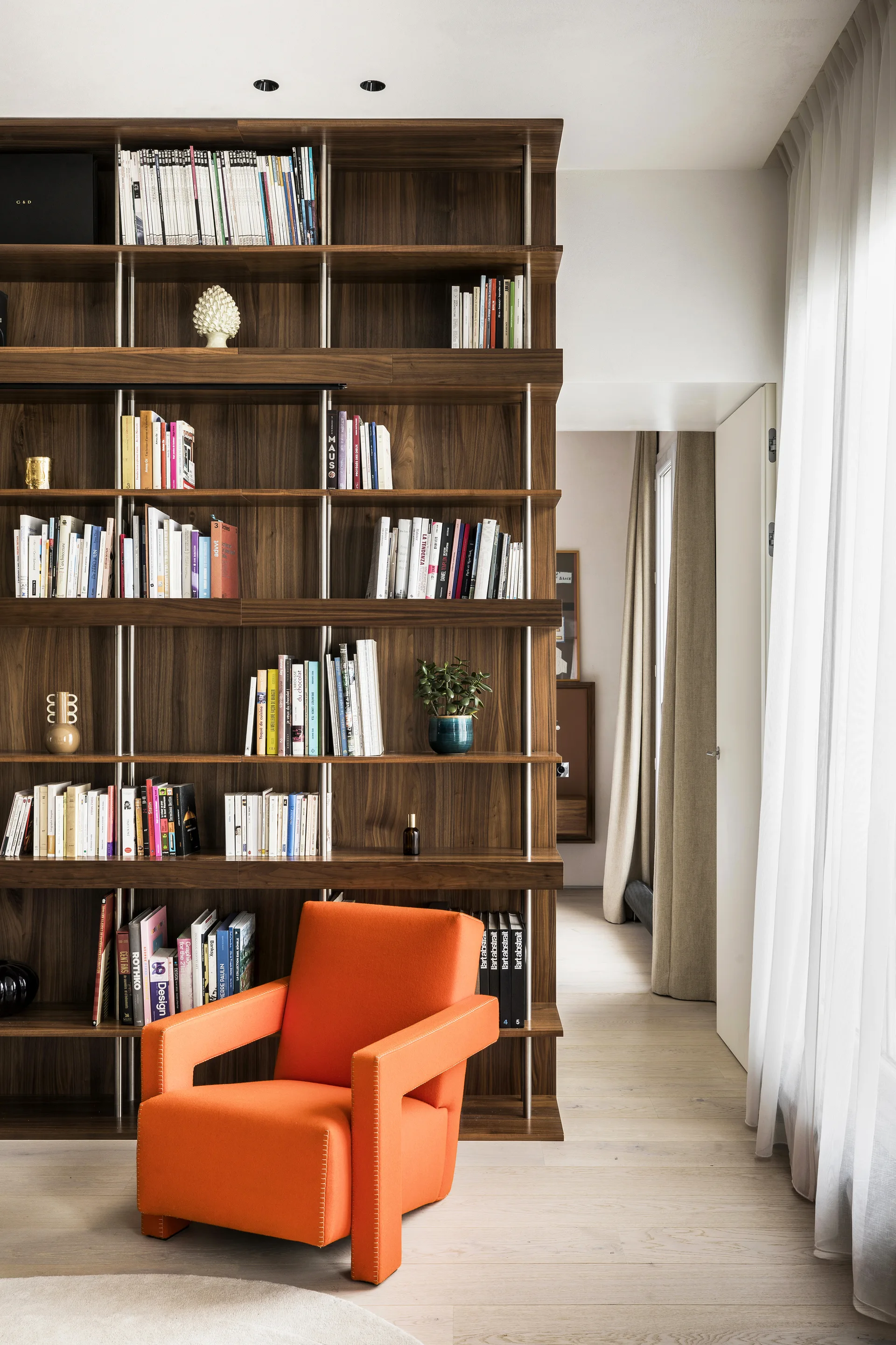 An orange armchair stands before a tall wood bookshelf with metal supports next to sheer white drapes.