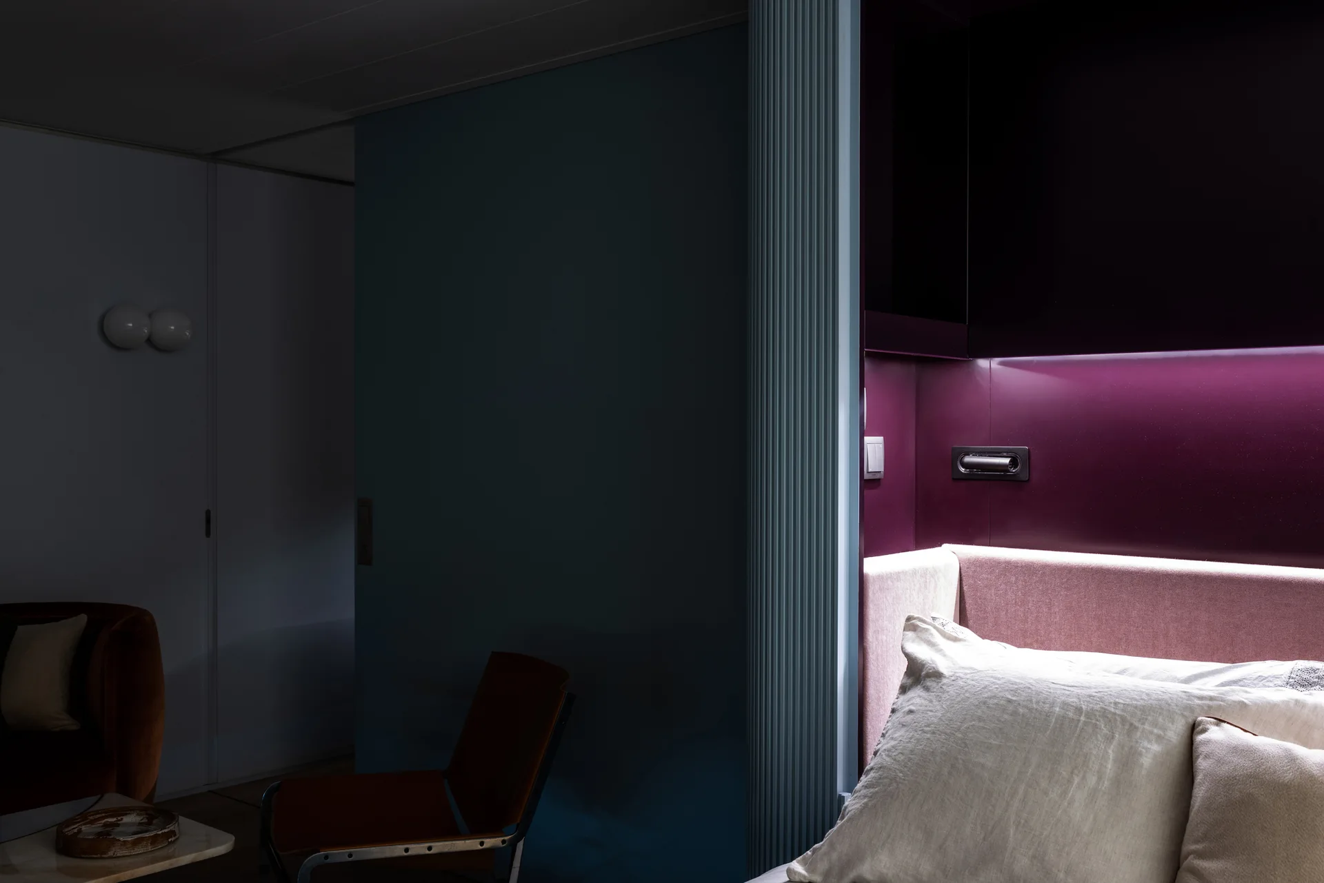 A contemporary bedroom corner featuring a pink headboard beneath an illuminated purple wall panel next to a light blue fluted column divider.