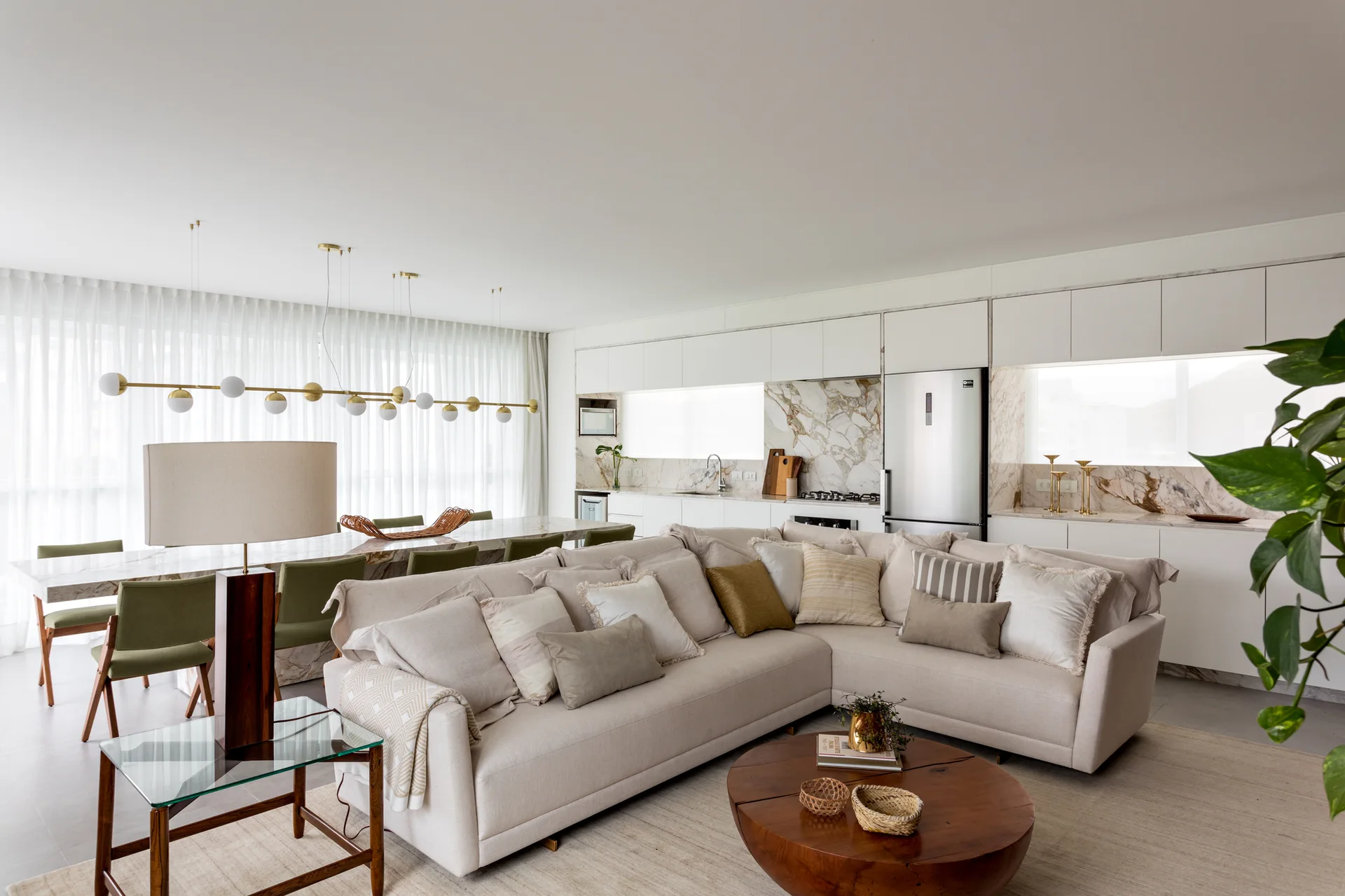 An open-concept living room with a cream sectional sofa, a round wooden coffee table, and an adjacent dining area featuring olive green chairs.