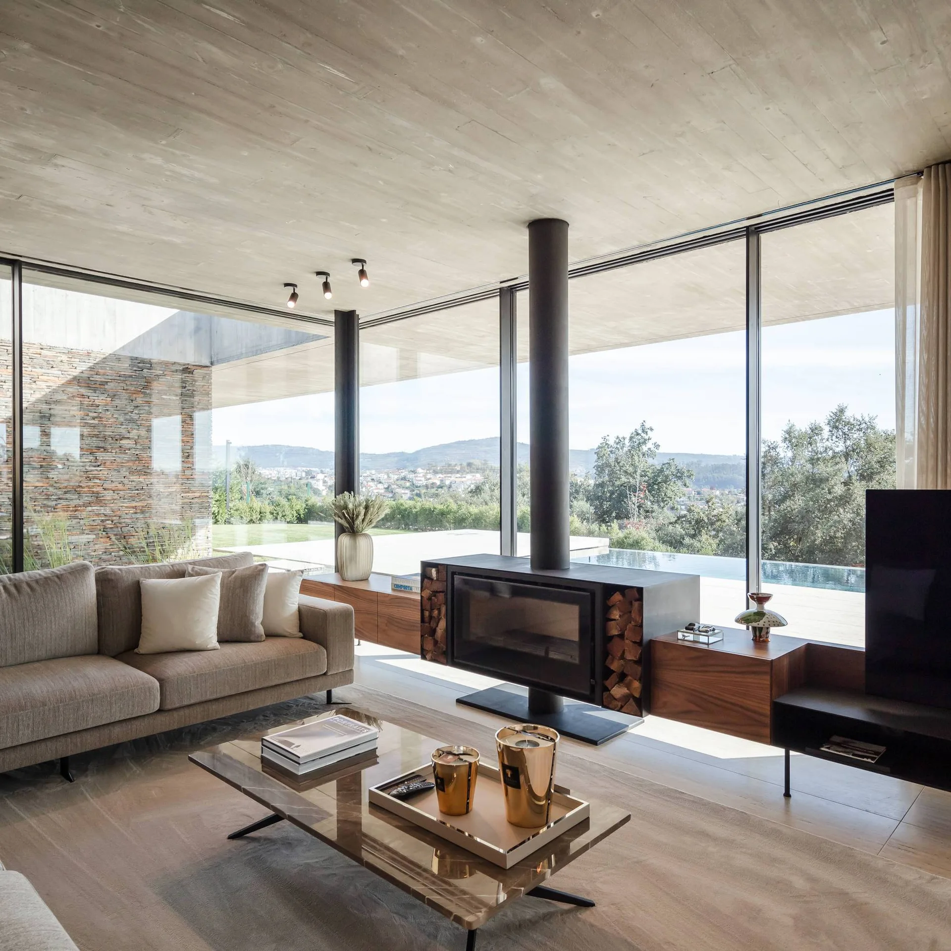 A modern living room features a beige sofa, a low stone coffee table, and a black freestanding fireplace in front of floor-to-ceiling glass walls.