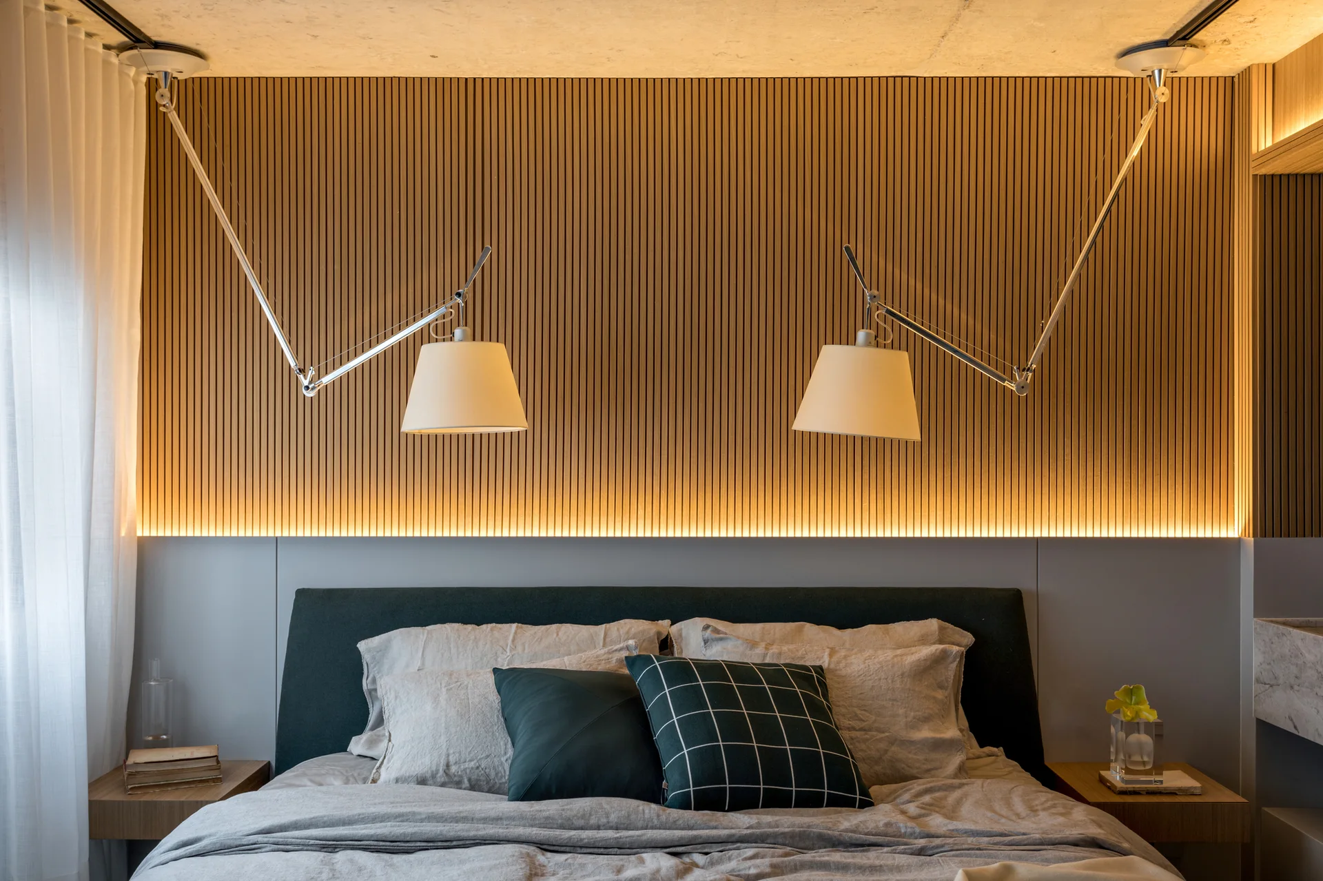 A modern bedroom features a vertical slatted wood accent wall with warm cove lighting above a dark upholstered headboard and twin swing-arm lamps.