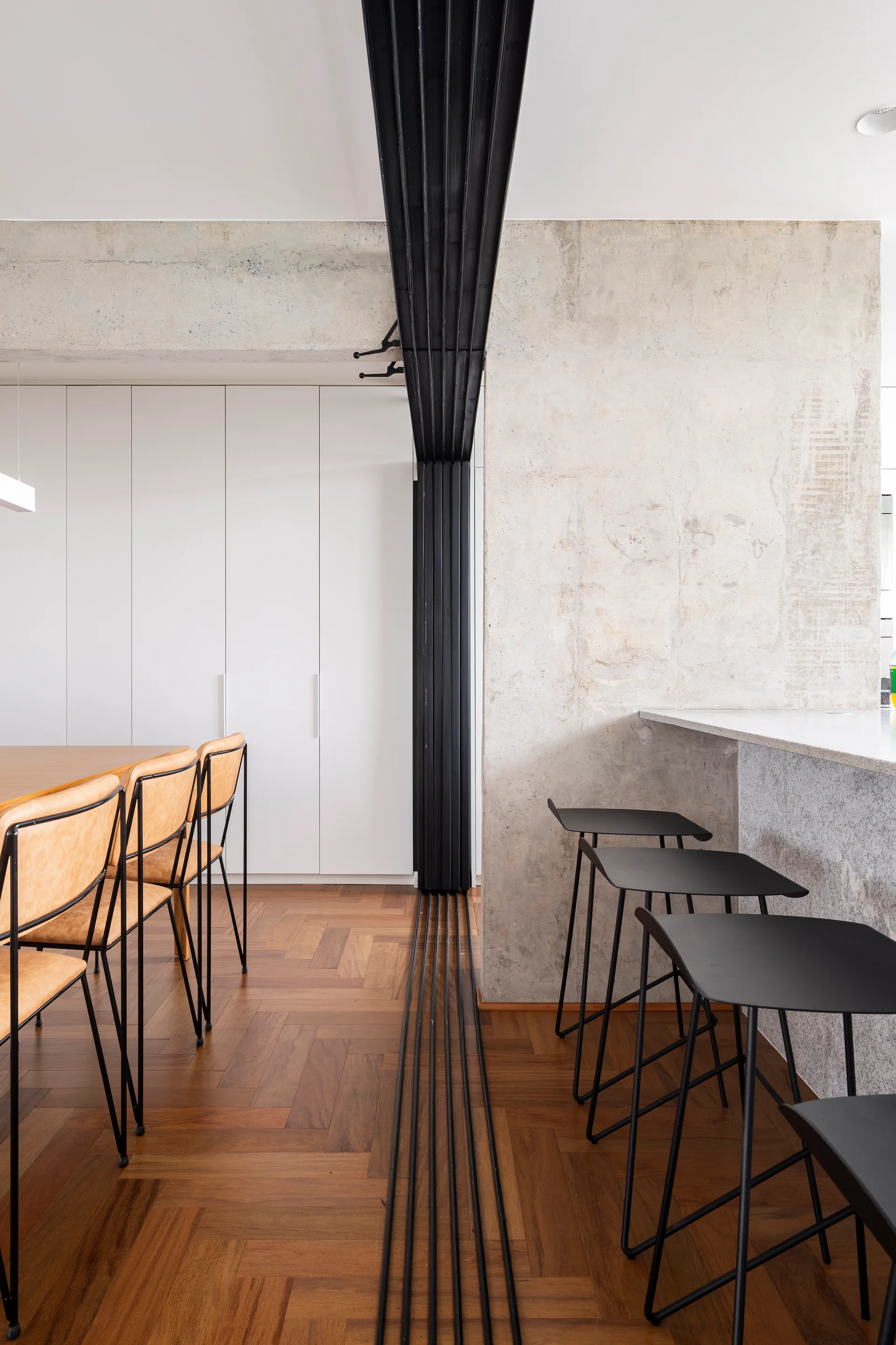 A modern dining space features tan leather chairs around a wood table, black metal bar stools at a concrete island, and a raw concrete accent wall.