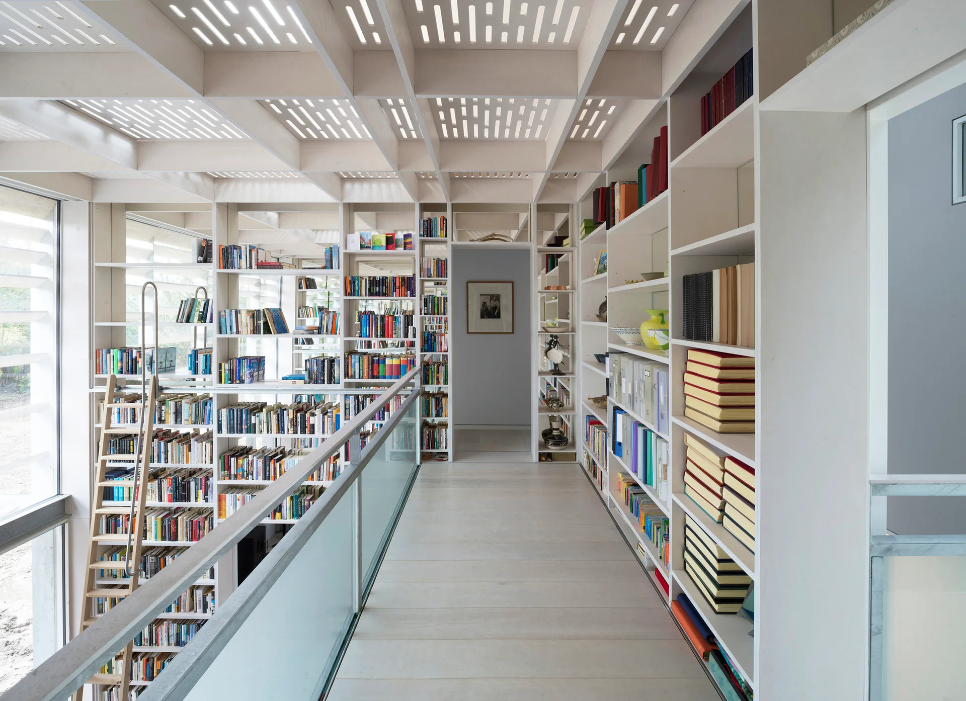 A modern library hallway features white floor-to-ceiling built-in bookshelves, a rolling ladder, glass railing, and a slotted wood grid ceiling.