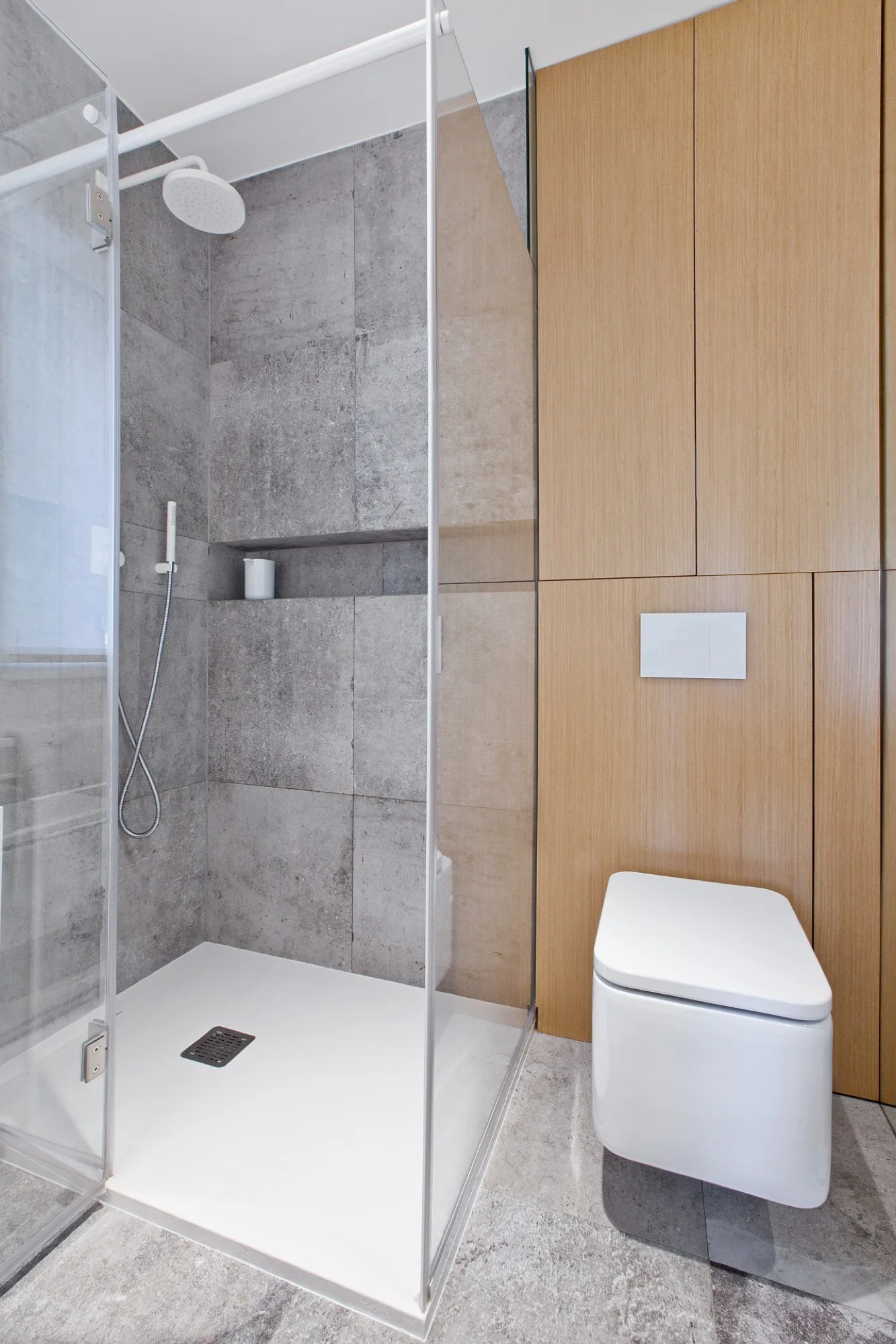A modern bathroom features a glass-enclosed walk-in shower with gray textured tile next to a wall-mounted toilet on a light wood paneled wall.