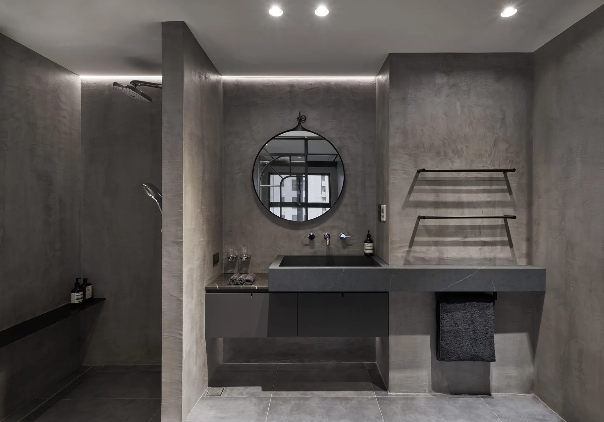 A minimalist grey bathroom features textured plaster walls, a floating vanity with a round mirror, and a walk-in shower behind a partition.