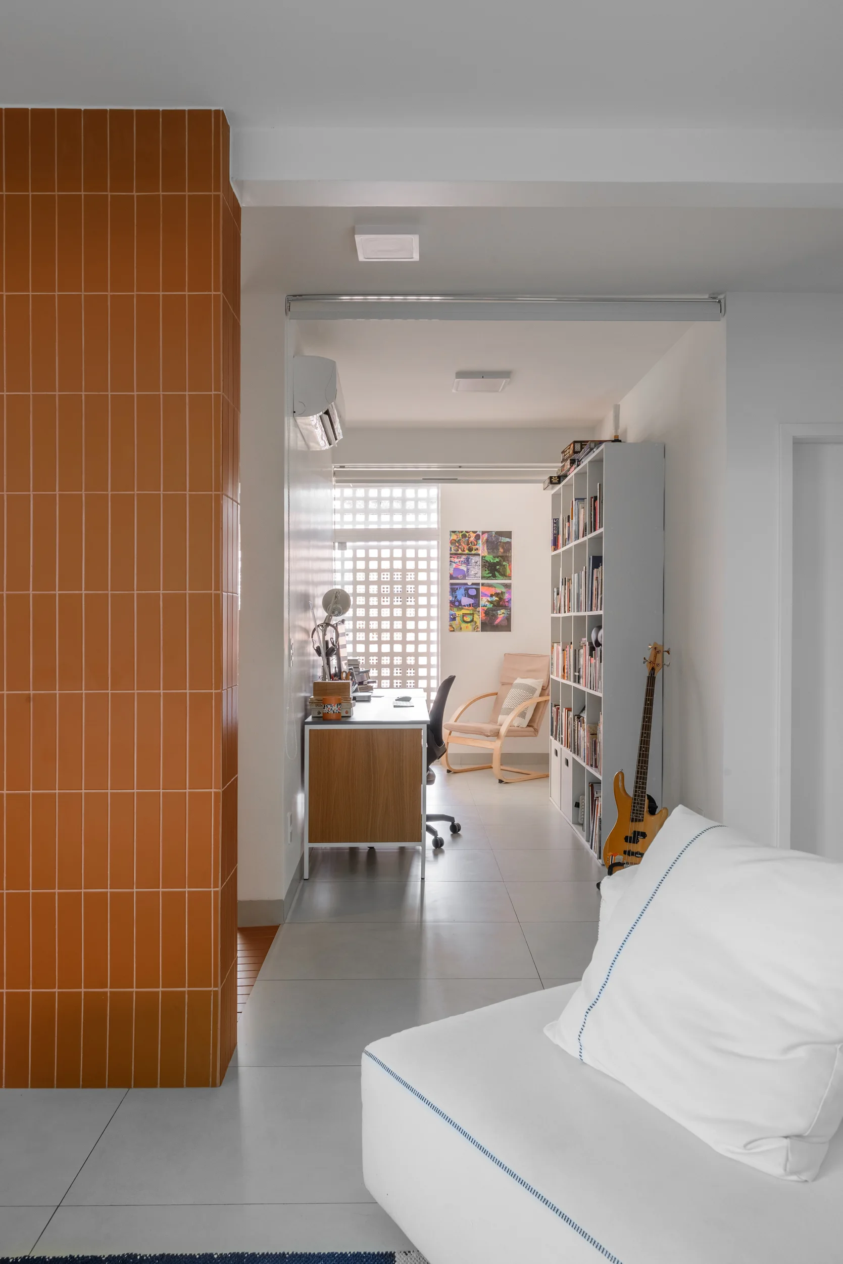 An interior view past a terracotta-tiled column into a bright home office featuring a desk, bookshelf, and a patterned breeze block window.