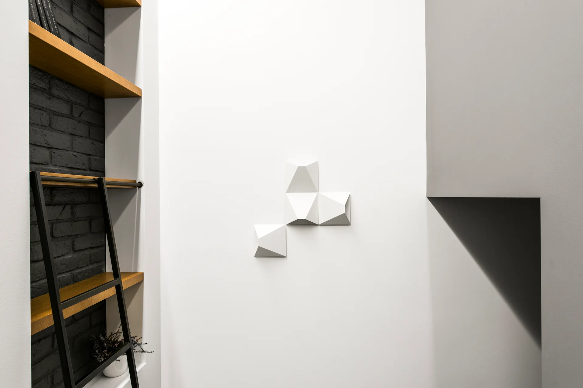A minimalist built-in wood shelving unit in a black brick alcove next to a smooth white wall featuring a geometric three-dimensional sculpture.
