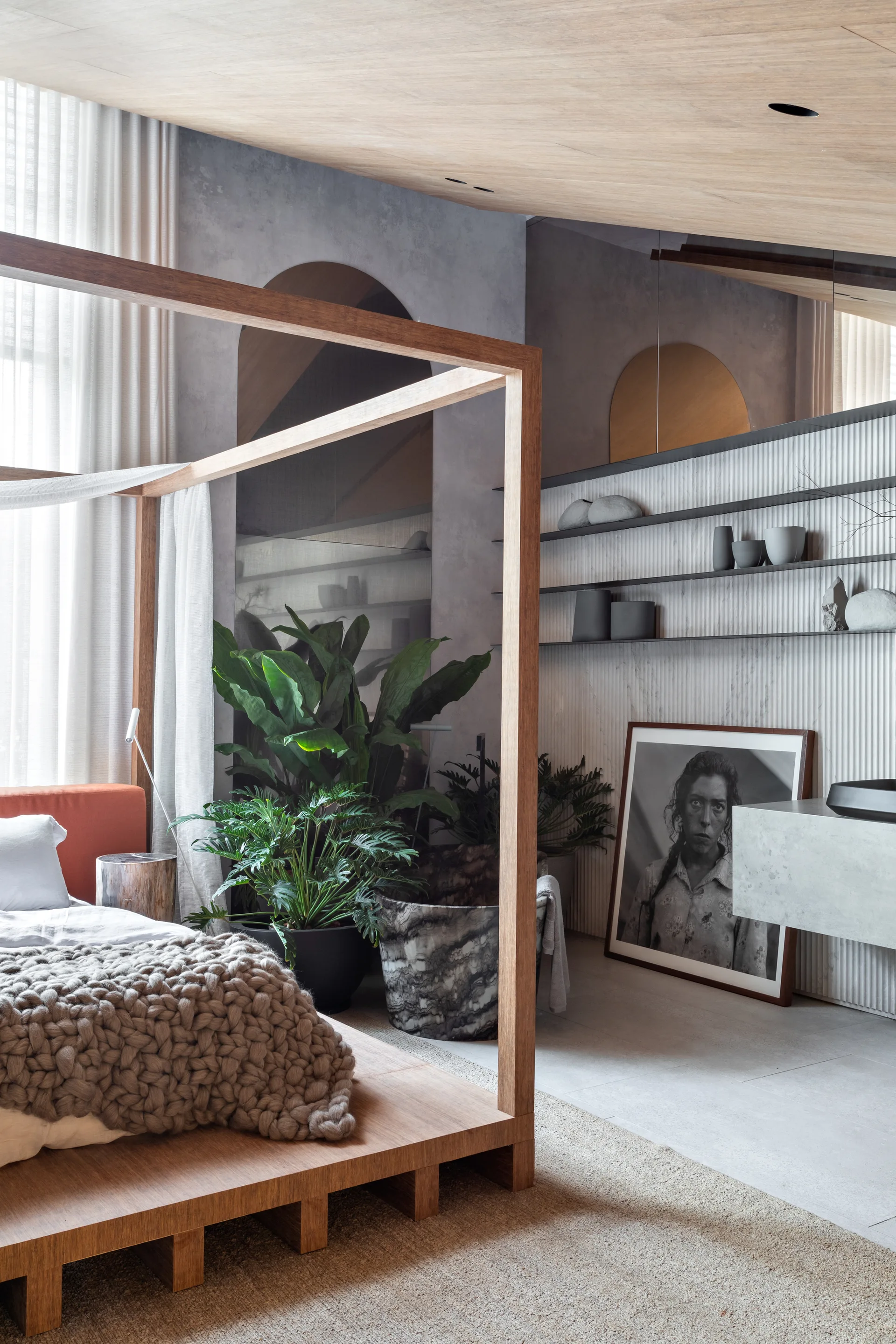 A wooden canopy bed sits in an organic modern bedroom featuring a fluted wall with floating shelves, large potted plants, and concrete finishes.