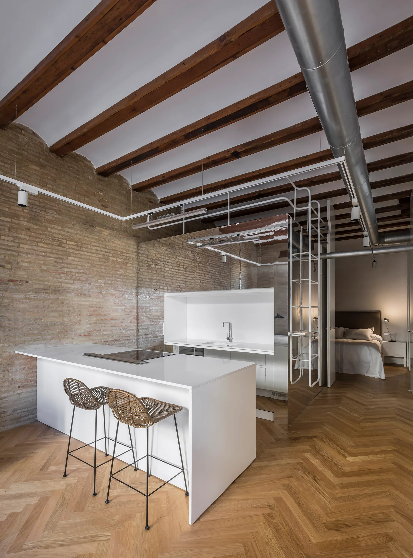 A modern kitchen island with rattan stools sits beside an exposed brick wall, timber ceiling beams, and a mirrored partition wall.