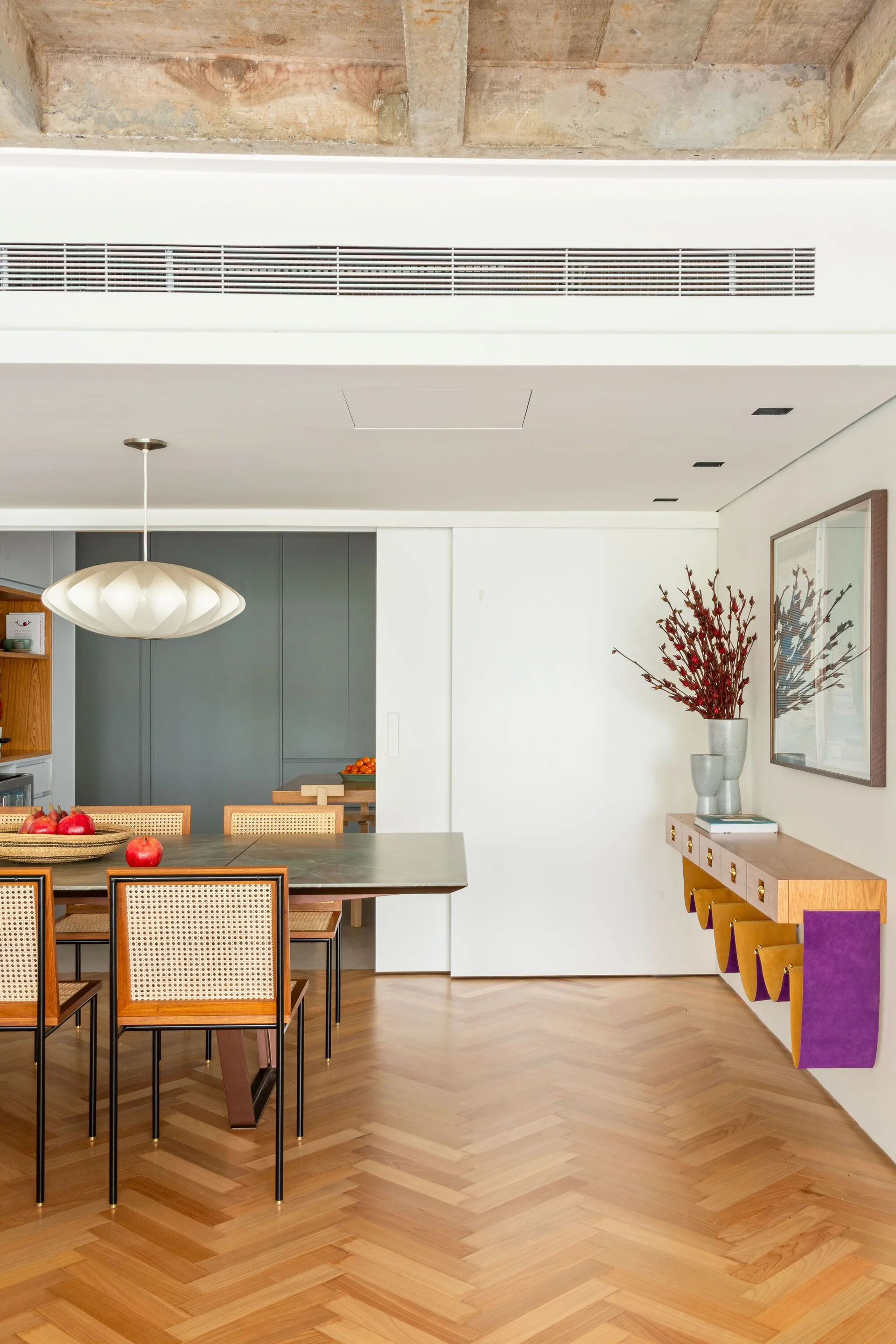 A modern dining room featuring cane-back chairs, a stone-top table, a floating console, herringbone wood floors, and an exposed concrete ceiling.
