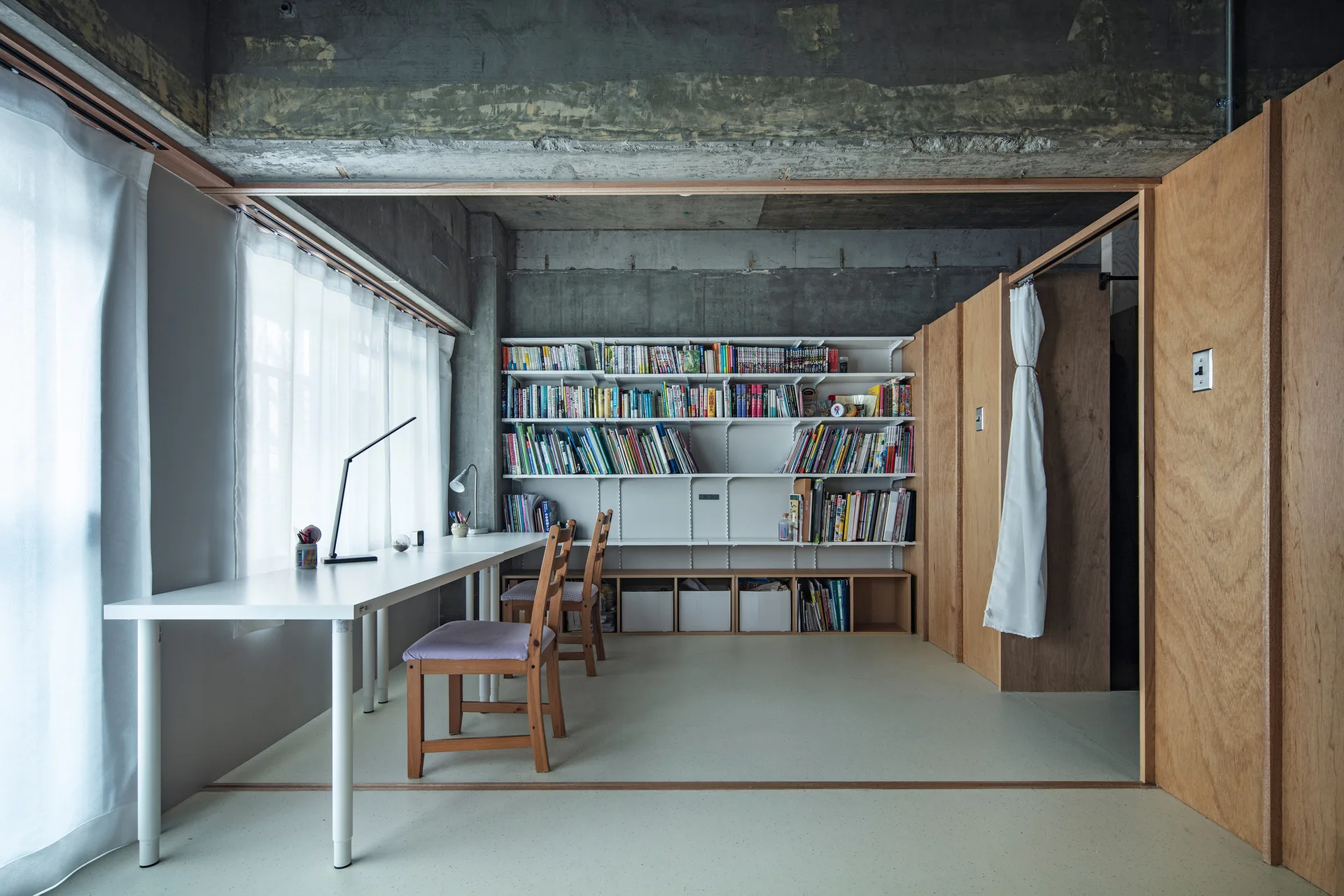 A minimalist home office featuring a long white desk, wall-mounted bookshelves, an exposed concrete ceiling, and plywood partition walls.